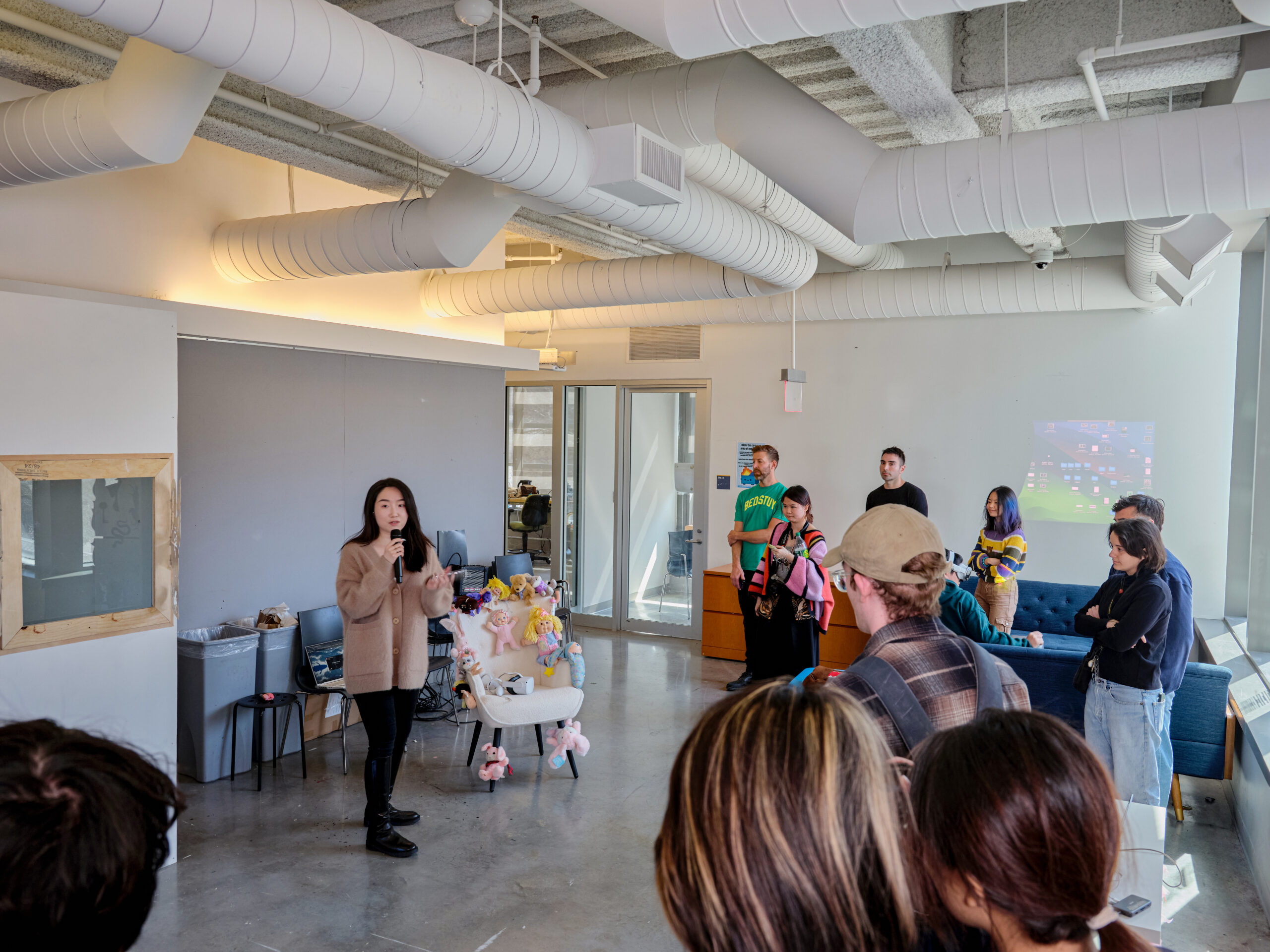 A student presents a project to an audience while speaking into a handheld microphone in a studio classroom. Attendees stand and sit around the space watching, with an installation featuring plush objects displayed beside the presenter and a projected computer desktop visible on the wall.
