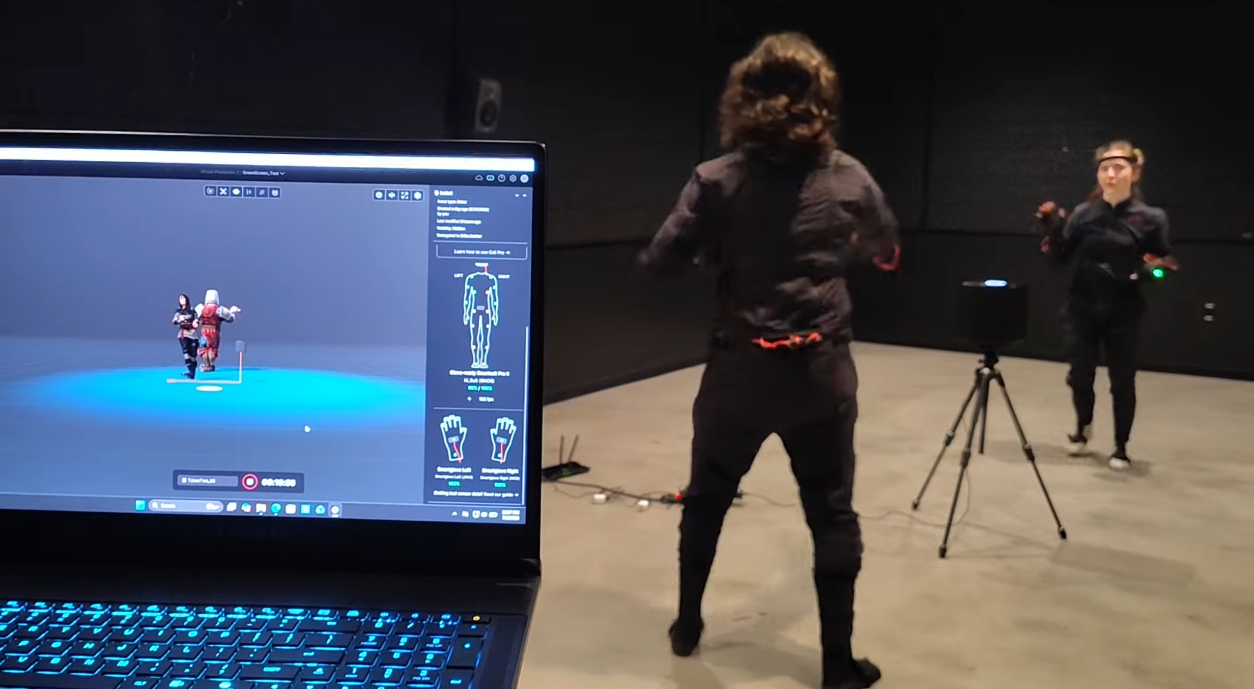 A student wearing a motion capture suit performs movement in a large studio while another student wearing a matching suit stands opposite. A laptop in the foreground displays real-time motion capture software with a live animated character preview, and a tracking camera on a tripod records the performers.