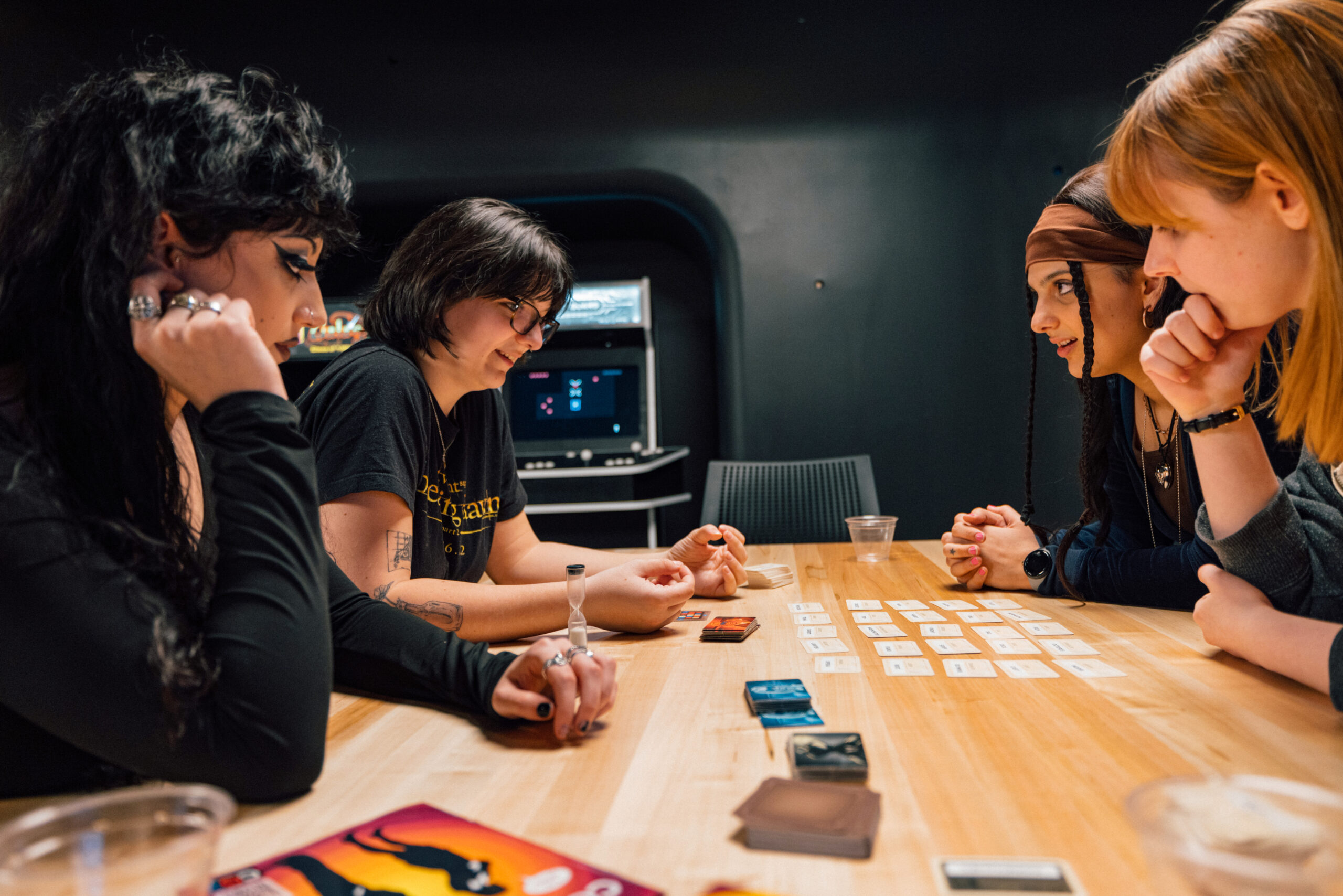Students and visitors participate in a game showcase inside a digital media lab filled with desktop gaming computers, monitors, keyboards, and illuminated computer towers. People gather around workstations playing games, observing demonstrations, and talking with developers while shelves of board games and presentation displays are visible throughout the space.
