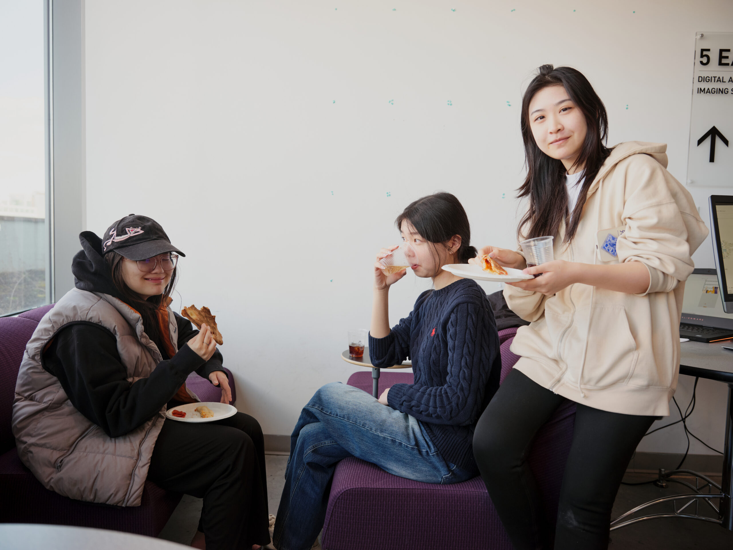 Three students take a break in a lounge area, eating pizza and drinking beverages while seated and standing beside a computer workstation. One student looks toward the camera holding a plate and cup as the others eat and converse.