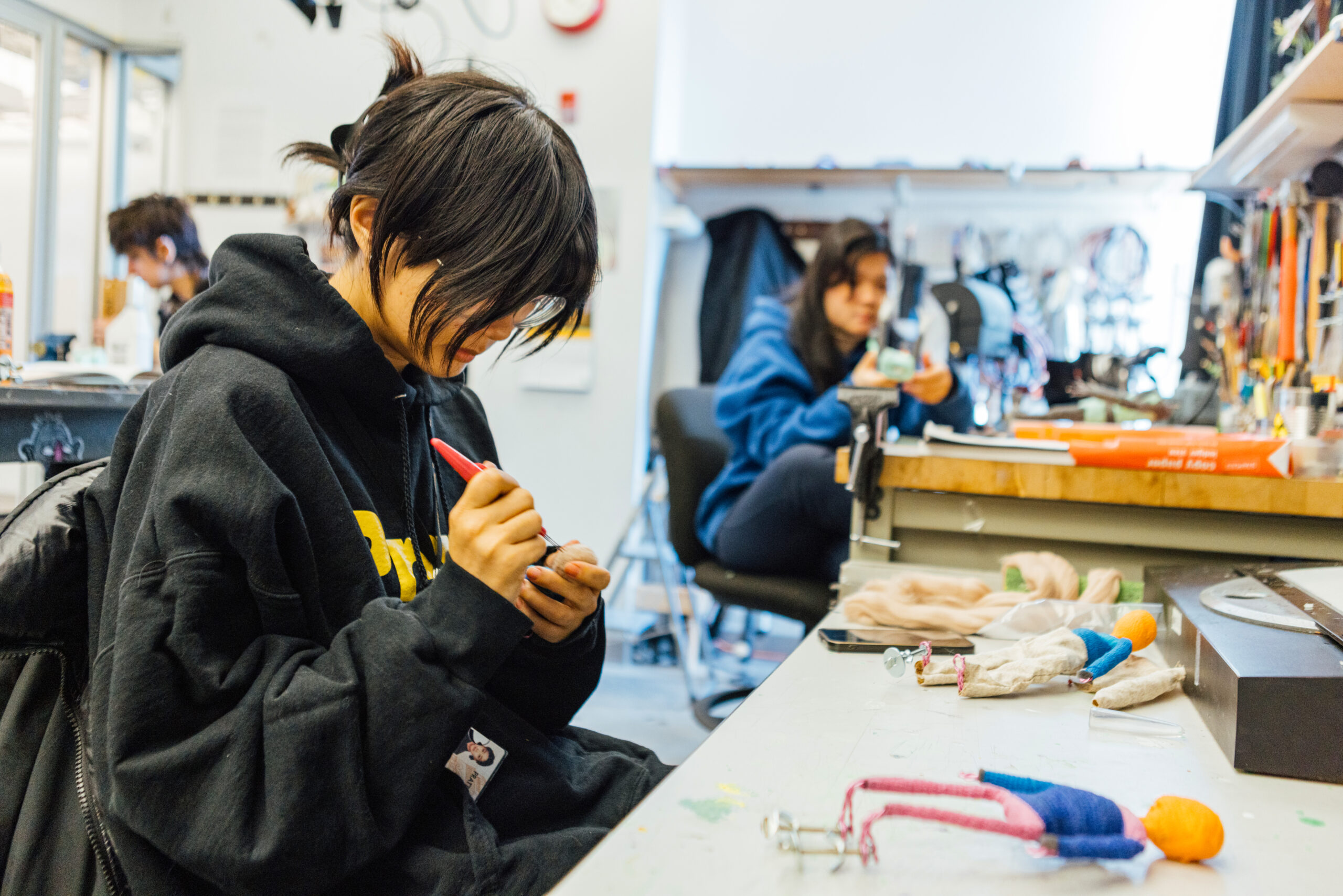 A student uses a sculpting tool to refine a small stop-motion puppet at a workbench covered with fabric, puppet parts, and animation materials, while another student works at a nearby station in the background.