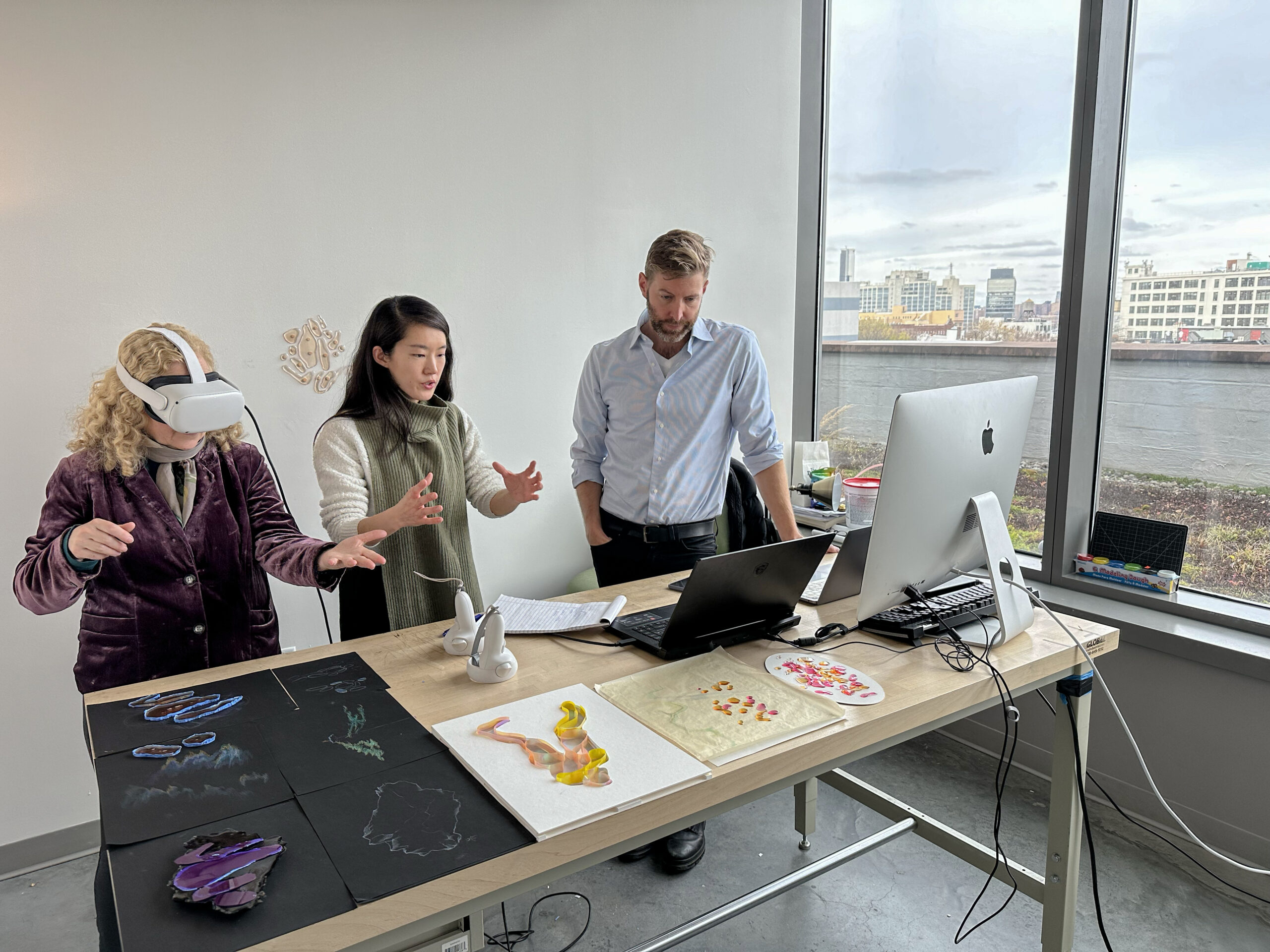 Three people gather around a workstation during a virtual reality project demonstration. One participant wears a VR headset and reaches forward with both hands while another gestures to explain the experience, as a third person observes beside a desktop computer. Prototype artwork and 3D-printed models are displayed across the table.
