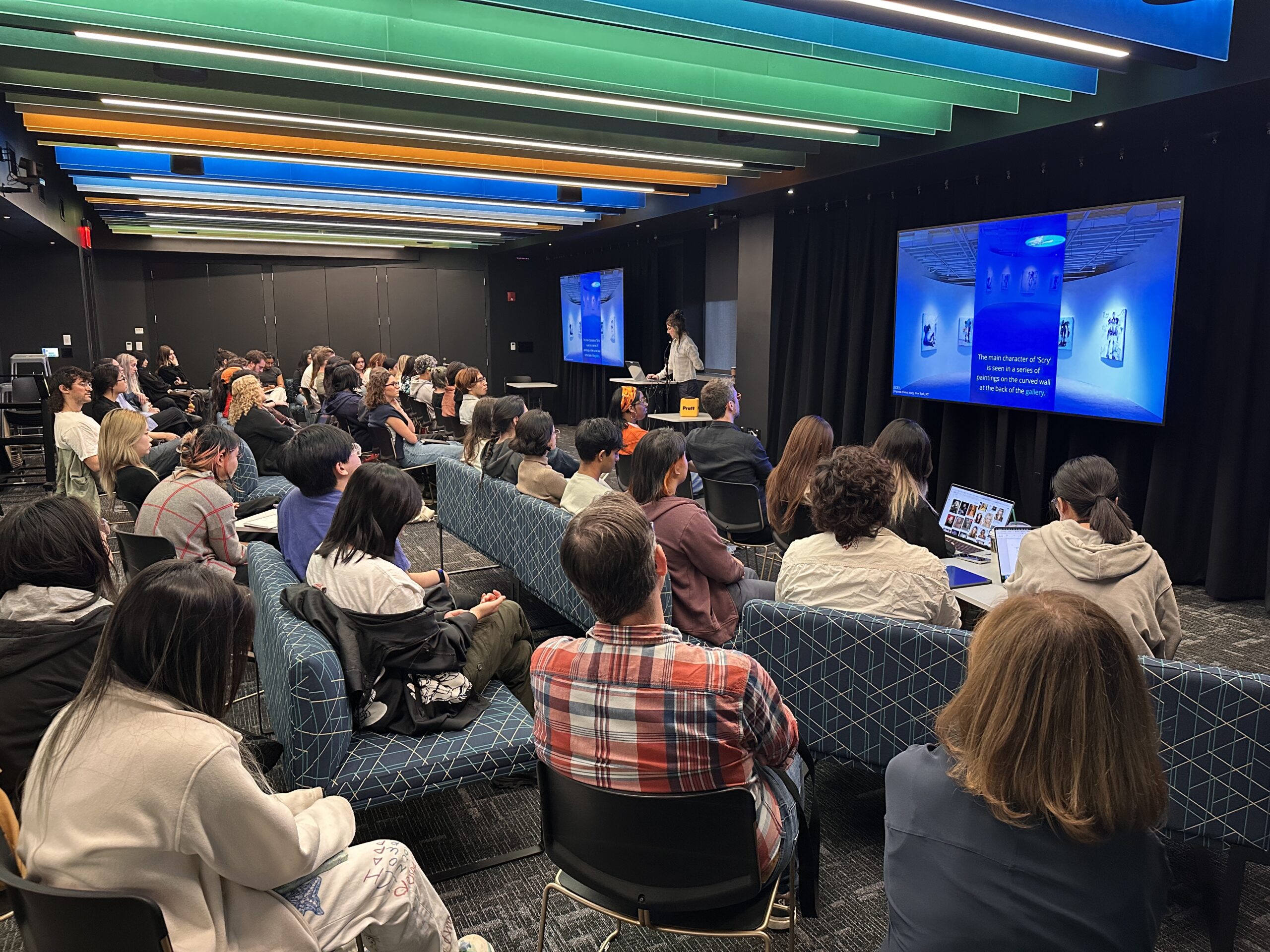 Audience seated in a modern presentation space attending a lecture while a presenter speaks beside large display screens showing digital artwork. Attendees sit on chairs and couches facing the front of the room beneath colorful illuminated ceiling panels, with several people taking notes or viewing laptops during the presentation.