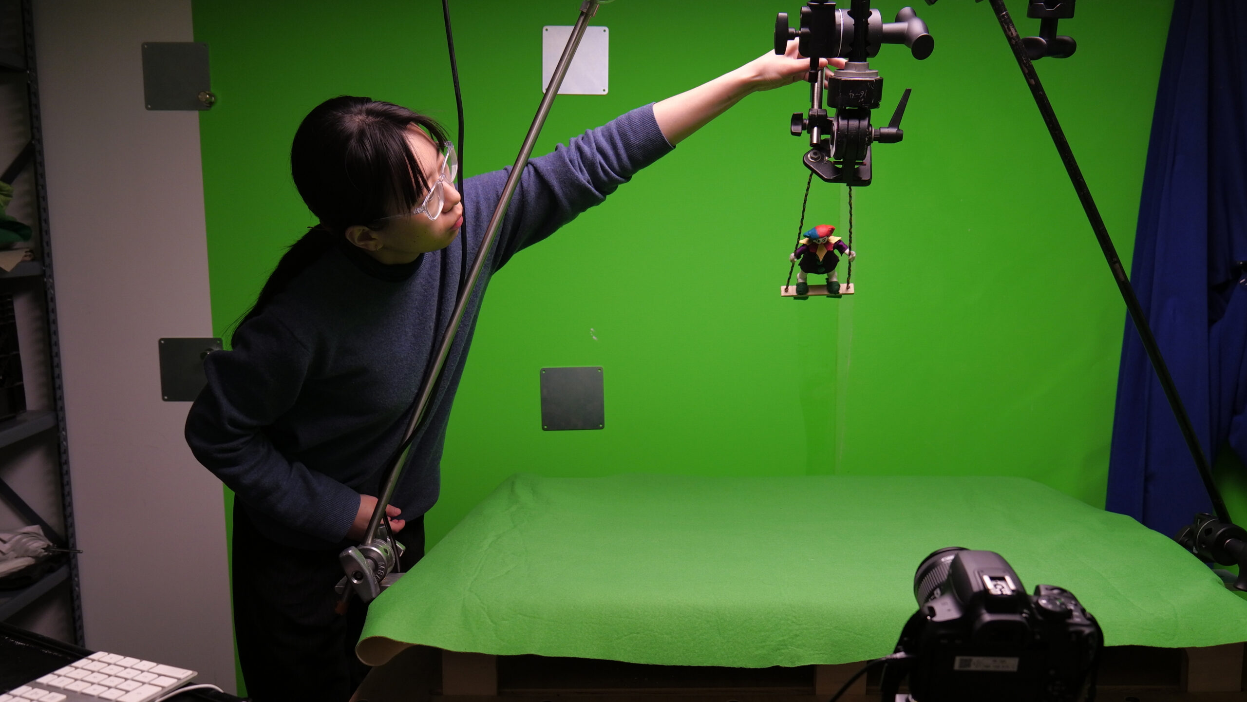 A student adjusts a camera rig suspending a small puppet on a swing above a green screen tabletop animation stage, with a DSLR camera positioned to capture stop-motion animation and studio equipment surrounding the workspace.