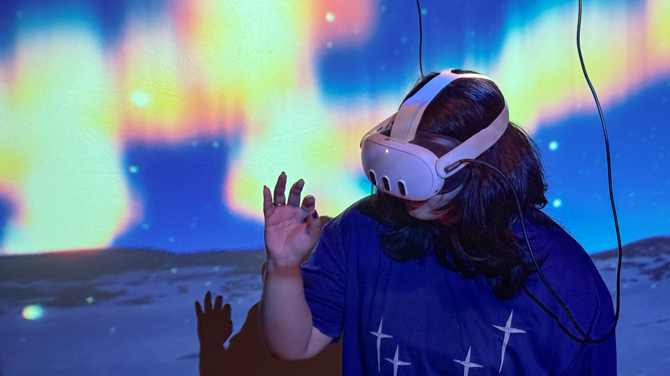 A person wearing a virtual reality headset gestures with one hand while immersed in a VR experience, with a colorful projection resembling the northern lights filling the background and the headset cable extending overhead.