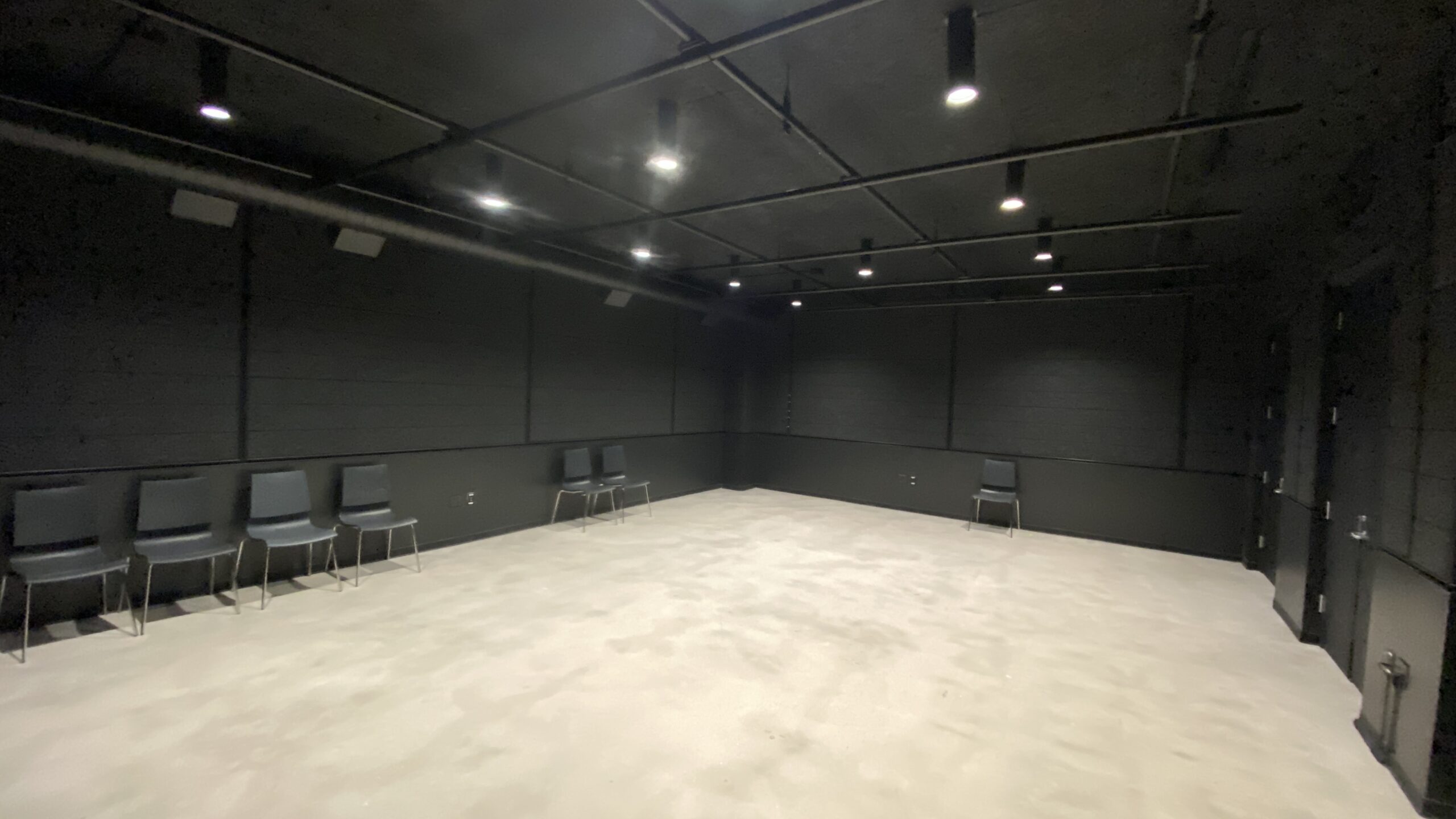 An empty black-box performance and rehearsal studio with black walls, overhead lighting, a polished floor, and several chairs arranged along the perimeter of the room.