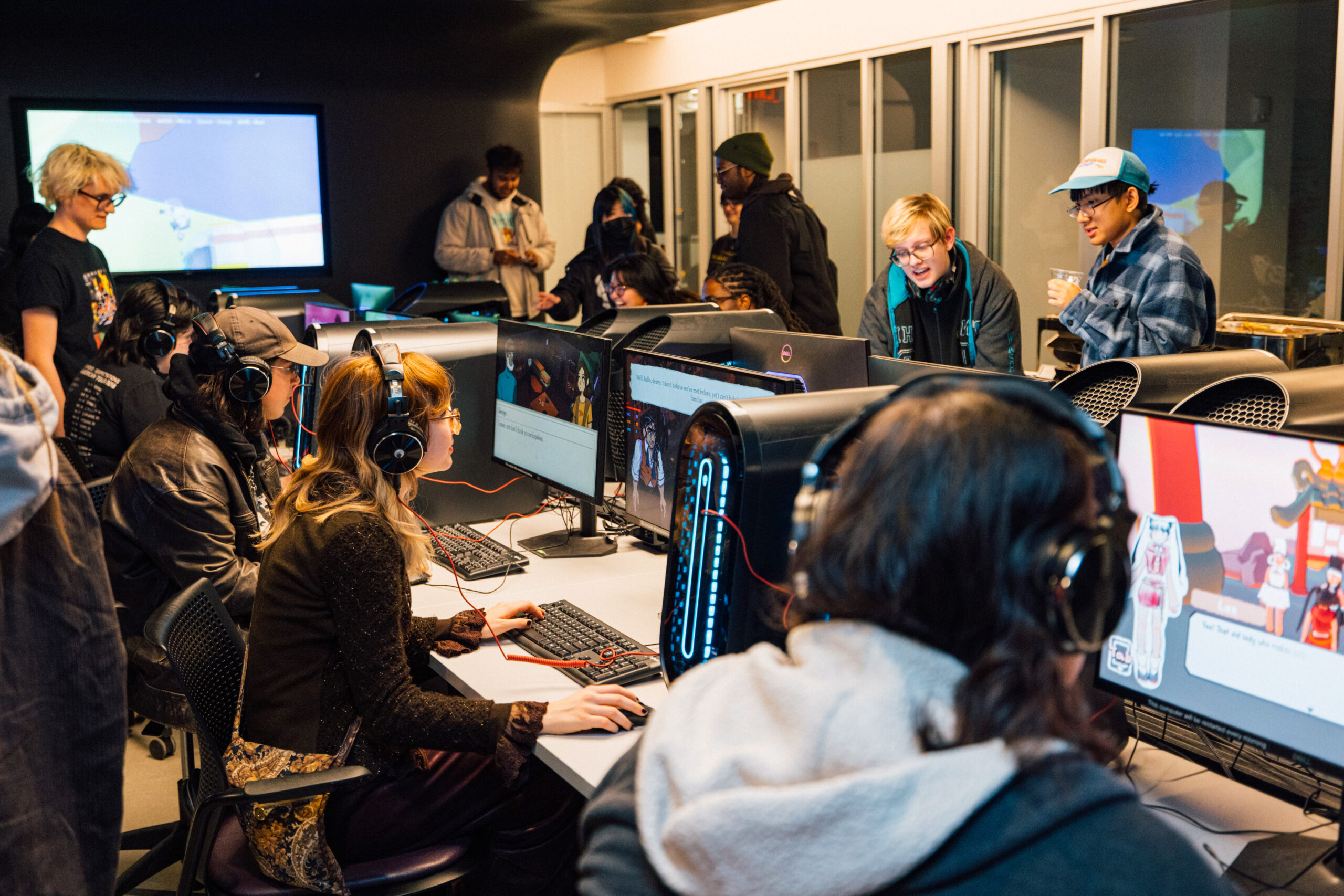 Student plays a computer game using a keyboard, mouse, and over-ear headphones at a desktop gaming workstation while others observe nearby. Multiple gaming computers with illuminated towers, monitors displaying gameplay, and attendees exploring student-created games are visible throughout the digital media lab.