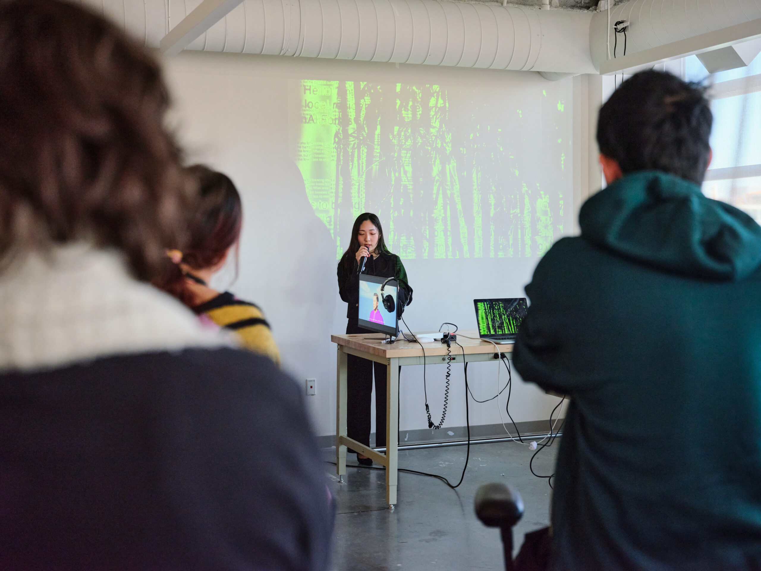 A student stands at the front of a room speaking into a microphone during a digital media presentation while audience members watch. A monitor, laptop, headphones, and projection displaying abstract green imagery are arranged on a table beside the presenter.