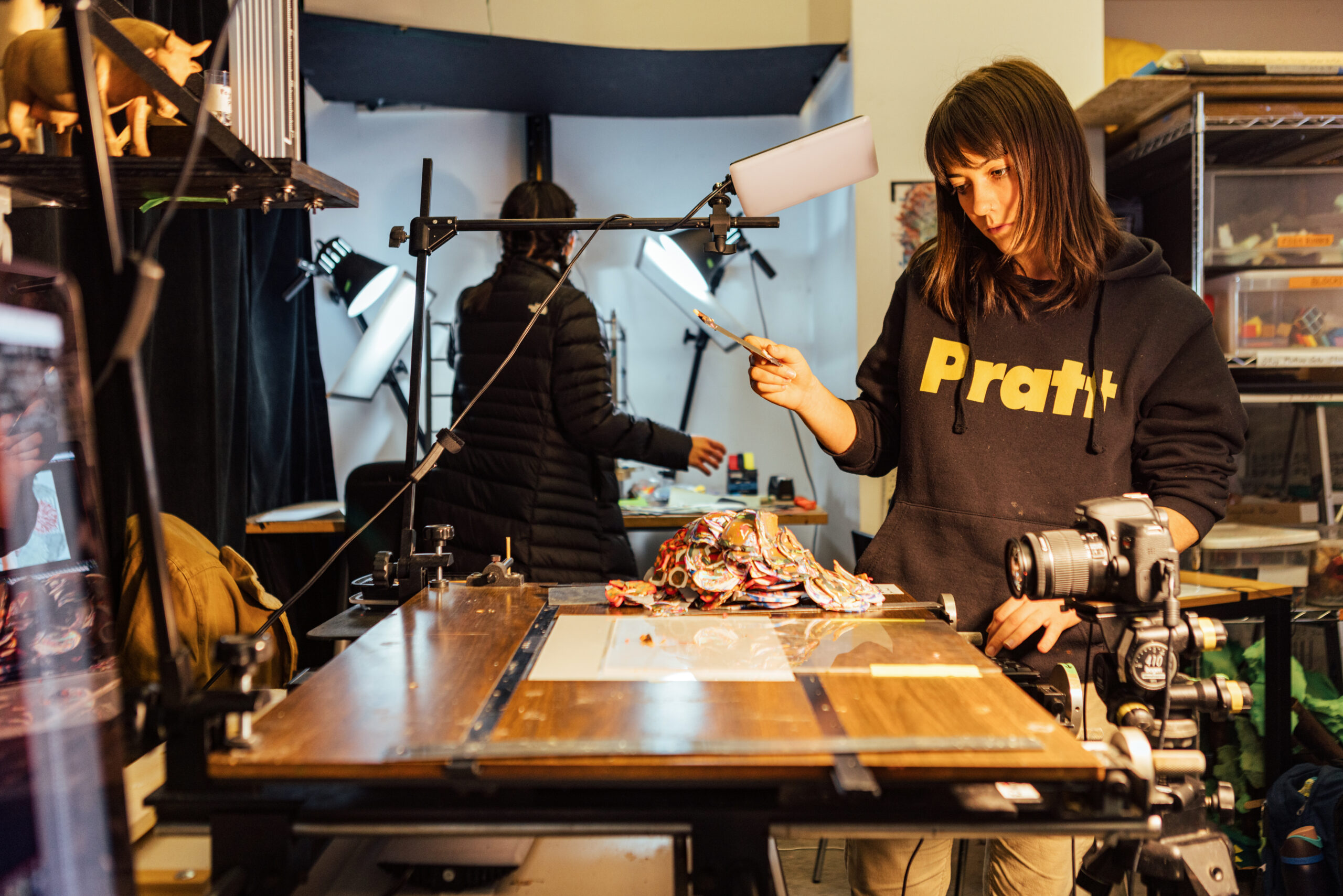 A student adjusts artwork on a stop-motion animation stand while holding a sculpting tool, with a DSLR camera mounted to capture frame-by-frame animation under studio lights in an experimental animation workspace.