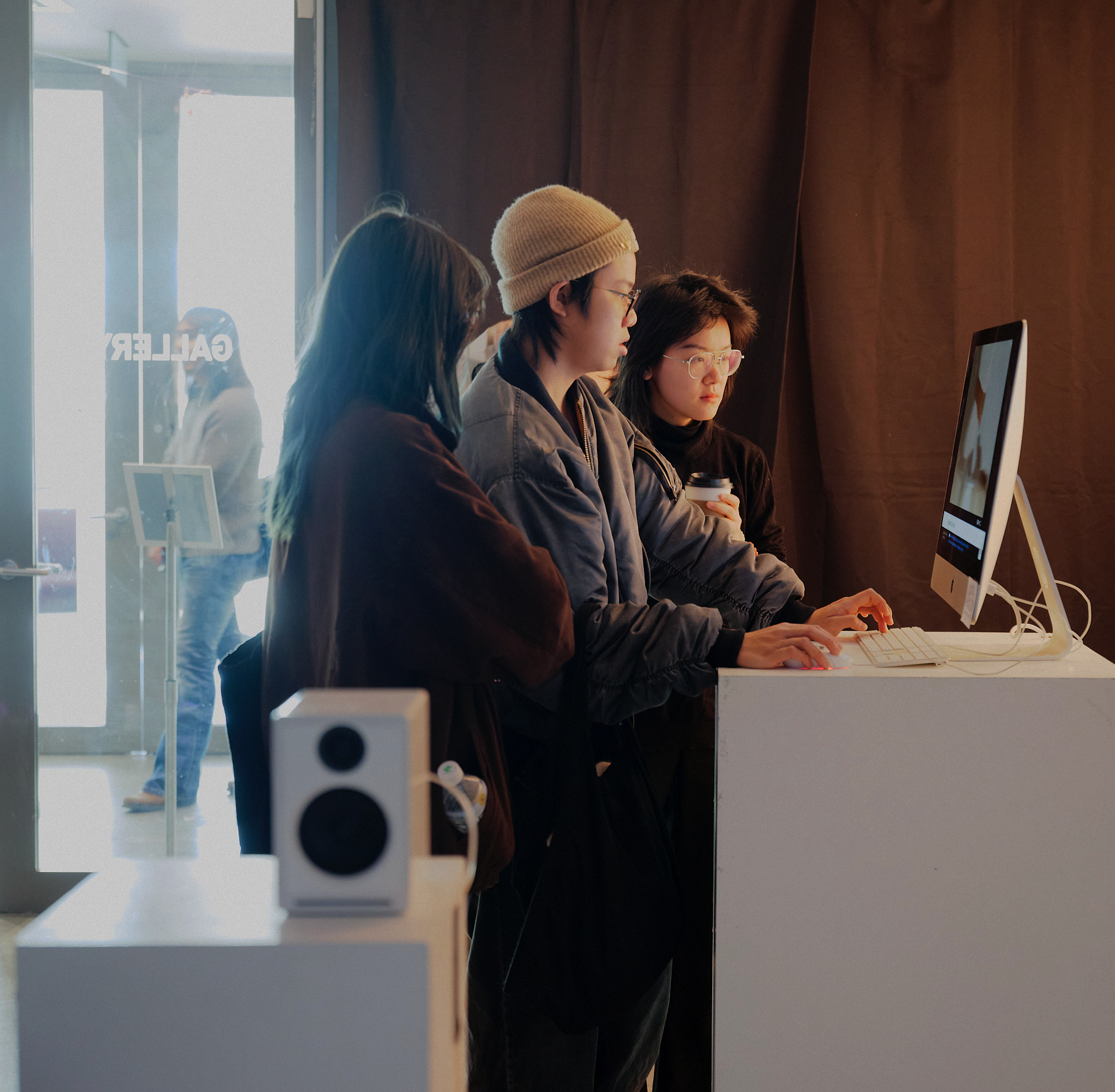 Two participants wearing virtual reality headsets stand in separate marked play areas on small green rugs while interacting with a VR experience. A white arcade-style display cabinet stands between them against a white gallery wall, with headset cables extending to the equipment and an open gallery entrance visible to the right.