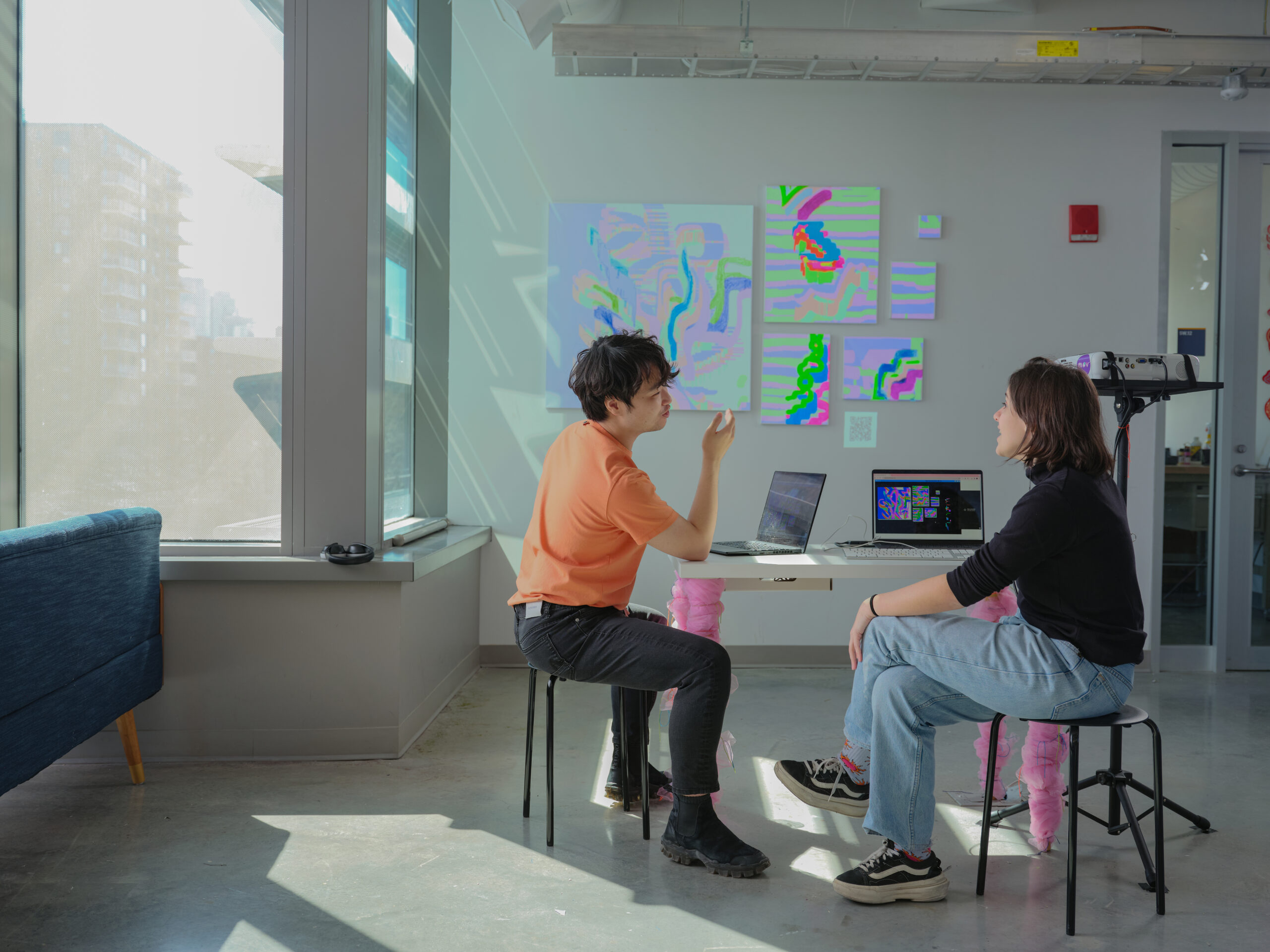 Two students sit across from each other discussing a digital art project at a table with two open laptops. Colorful abstract digital artwork is projected onto the wall behind them, and a projector stands nearby in a bright studio space.