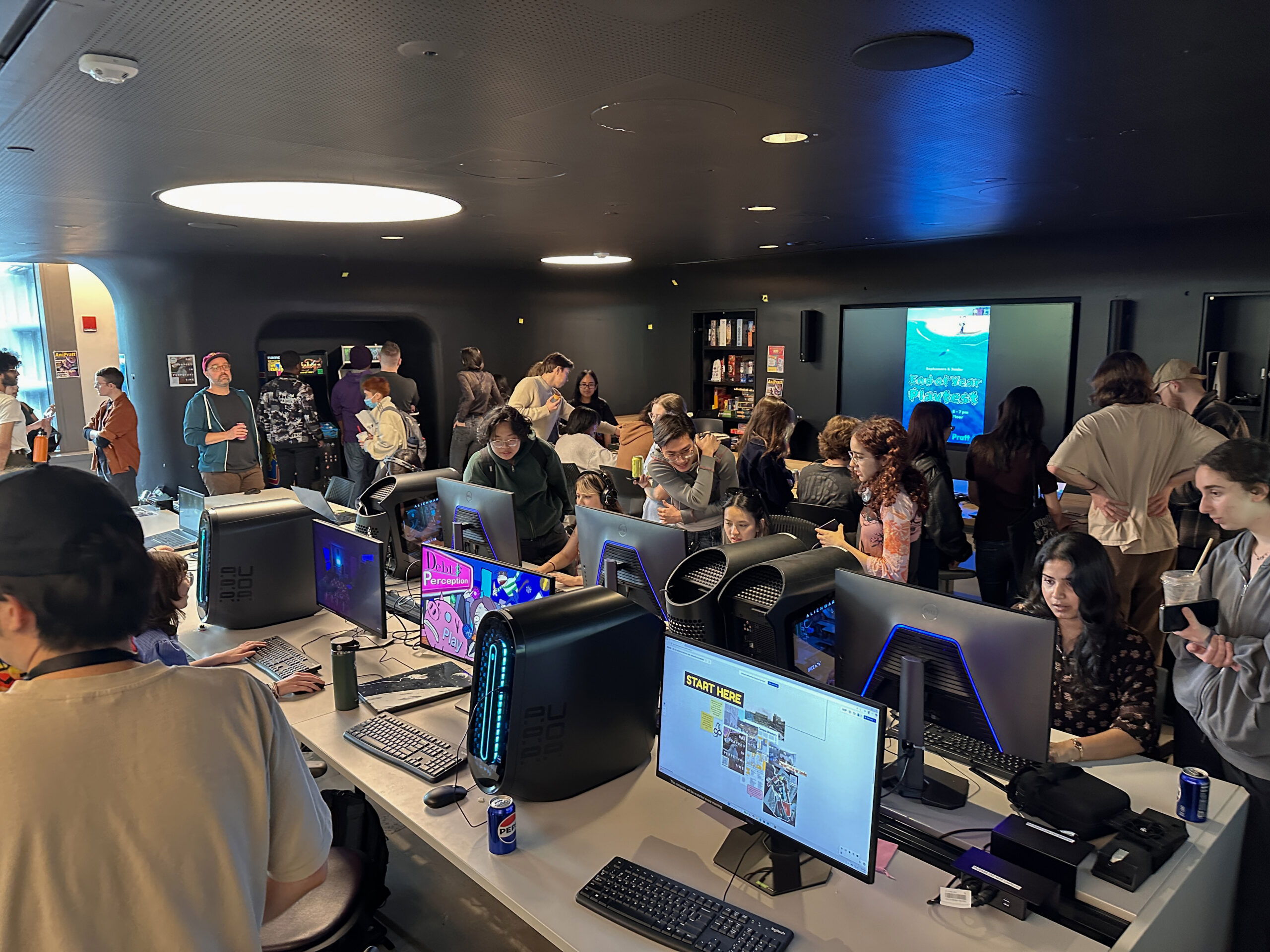 Students gather in a digital media lab for a social event, talking in small groups around a large communal table while others use computer workstations in the background. Plates, refreshments, board games, arcade cabinets, and desktop computers create a shared space for community building, collaboration, and informal interaction.
