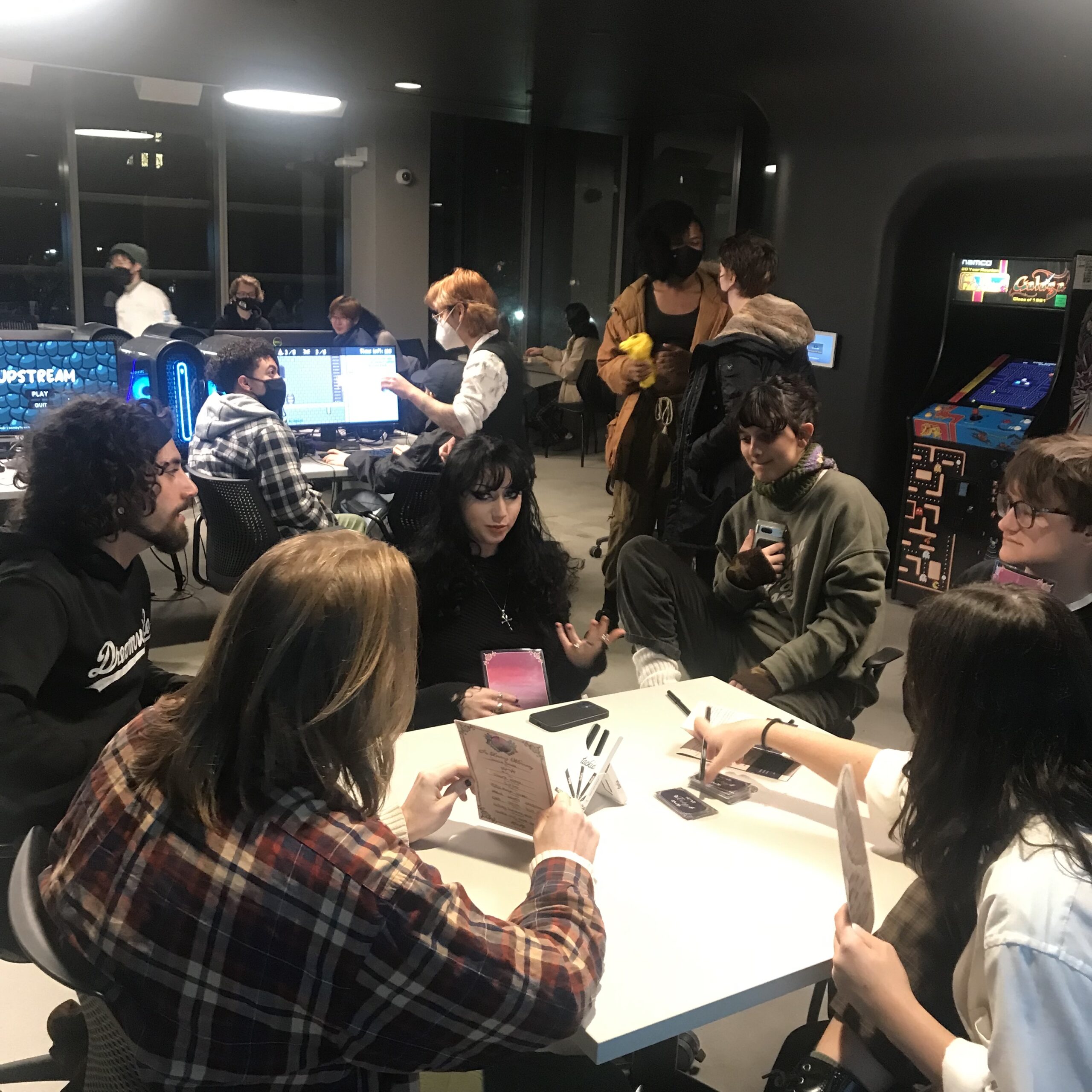 Students and visitors play multiplayer computer games at rows of desktop gaming workstations equipped with monitors, keyboards, mice, over-ear headphones, and illuminated computer towers. Others stand nearby watching gameplay, offering feedback, and socializing while arcade cabinets and additional gaming stations are visible in the background.