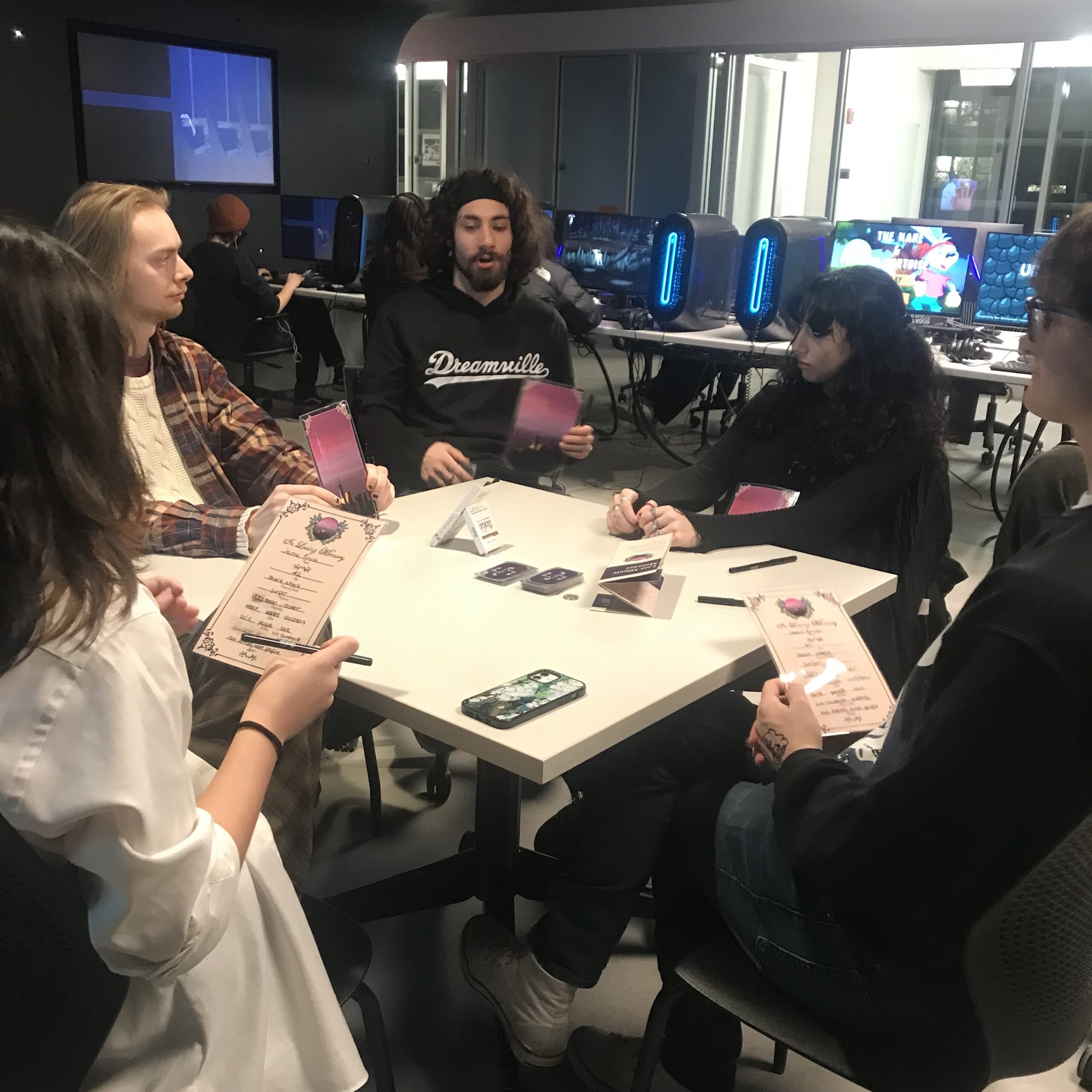 Students participate in a collaborative gaming event using desktop gaming computers equipped with monitors, keyboards, mice, and illuminated computer towers while others observe gameplay and wait their turn. Groups of participants gather around the room socializing, discussing games, and exploring the digital media lab's gaming and arcade spaces.
