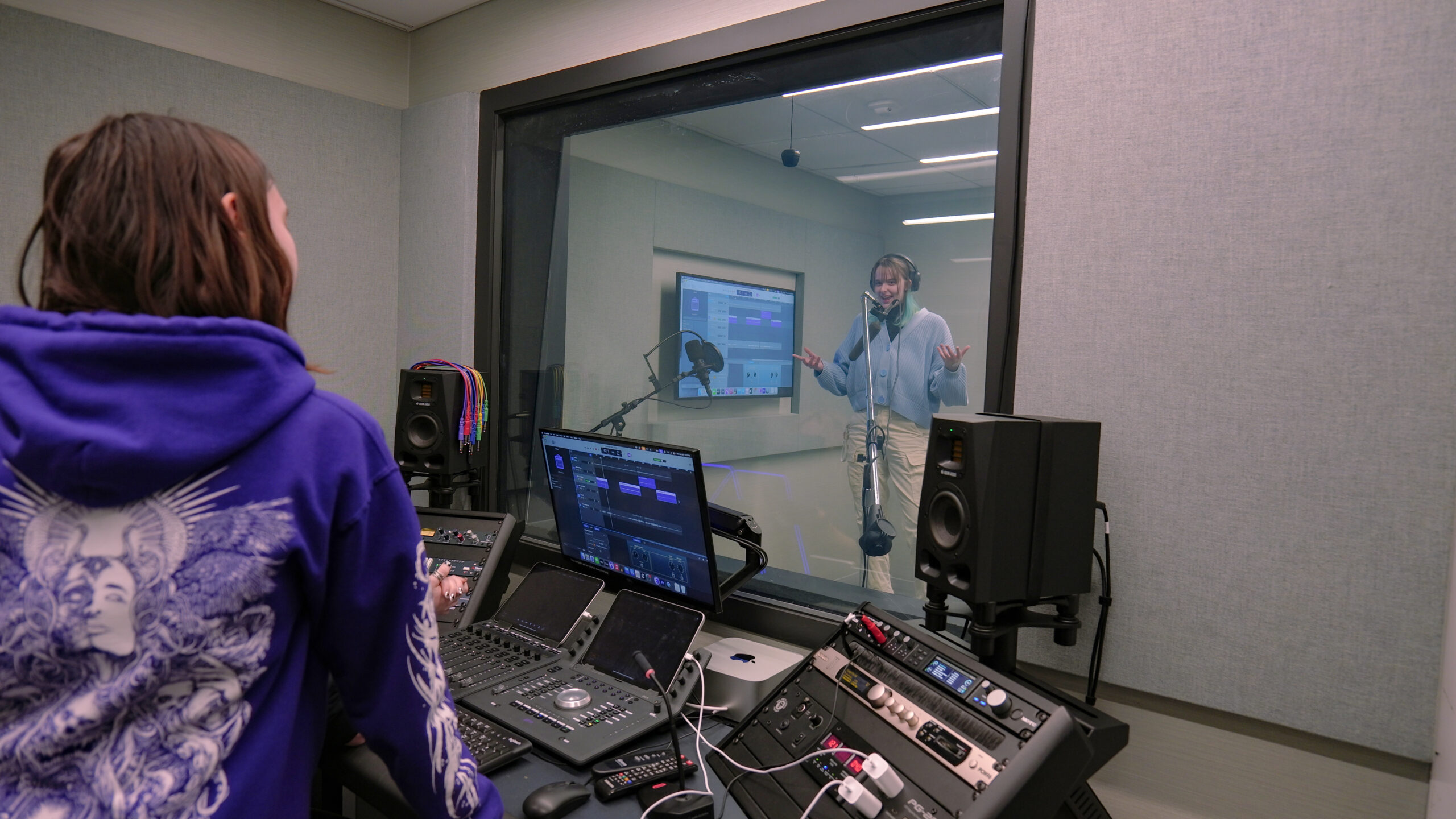 Students collaborate in a professional audio recording studio as one student performs in a vocal booth wearing headphones and speaking into a studio microphone while two classmates monitor and direct the recording from the control room. Digital audio workstation software, studio monitor speakers, audio interfaces, and mixing equipment are used to review and refine the recording session through the soundproof glass window.