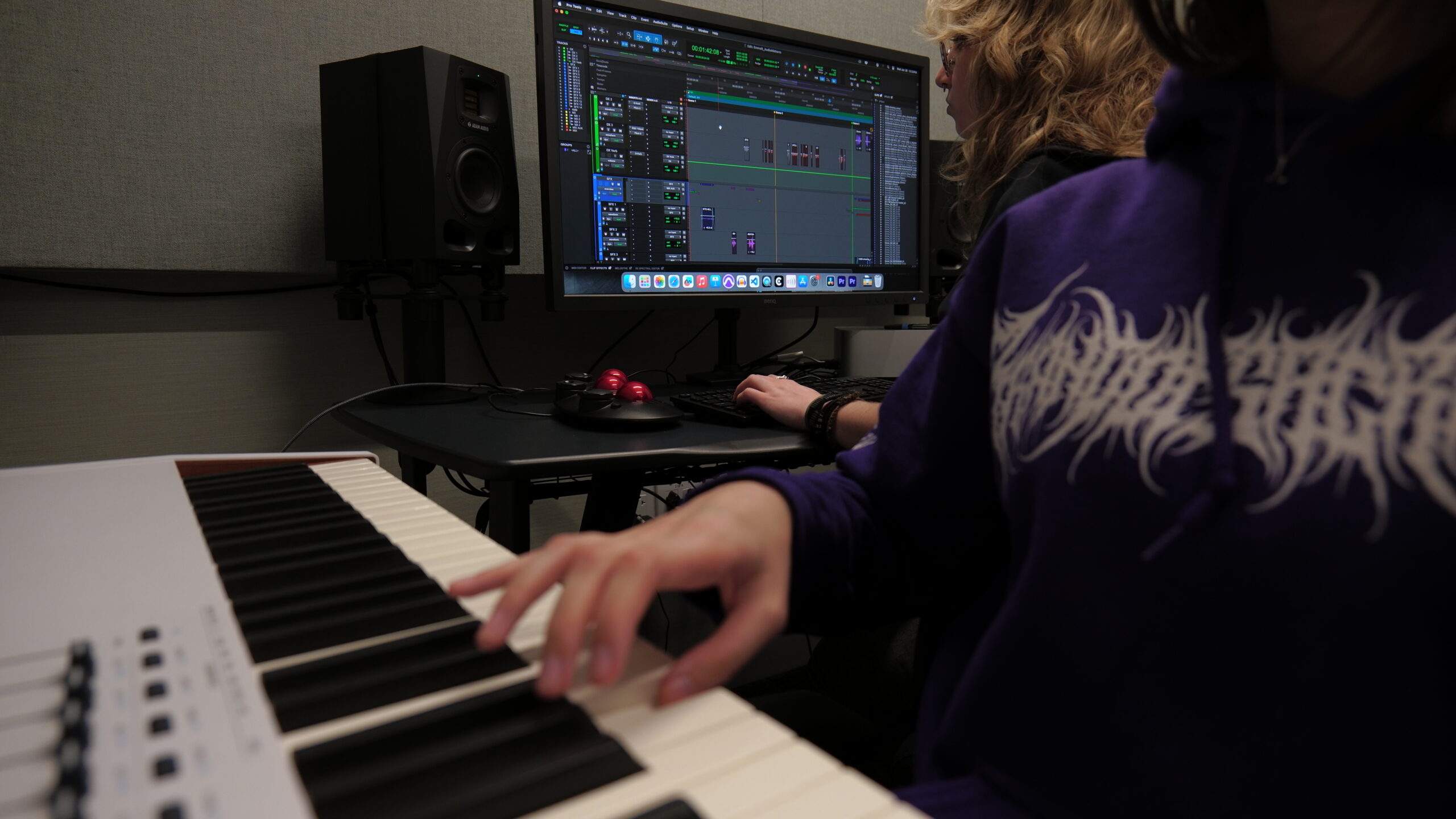 Student plays a MIDI keyboard while another student edits a multitrack audio project in professional digital audio workstation software on a desktop computer inside an acoustically treated recording studio. Studio monitor speakers, editing controls, and music production equipment surround the workstation as the pair collaborate on recording and audio editing.