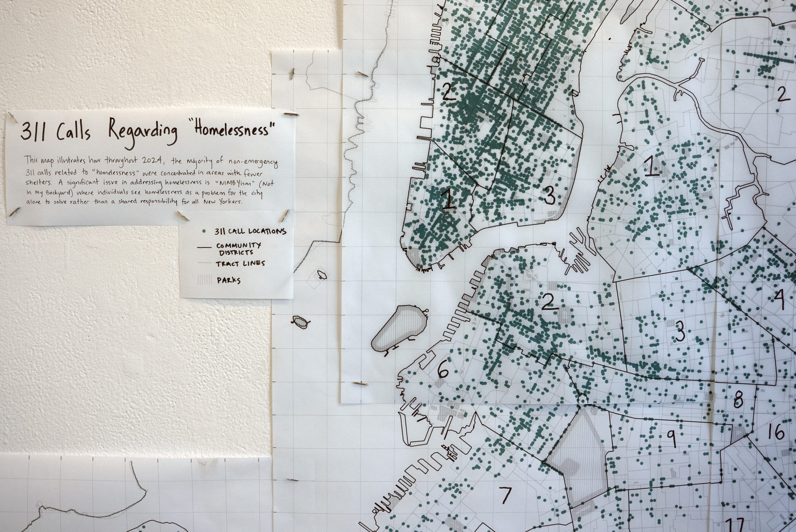 A map shows the locations of 311 calls regarding homelessness from 2024, marked by green dots. An accompanying sign explains the focus on areas with fewer shelters and discusses the issue of NIMBYism (Not In My Backyard). The map features outlined community districts, tract lines, and parks, with some sections labeled numerically. The overall context highlights a shared responsibility for addressing homelessness in New York City.