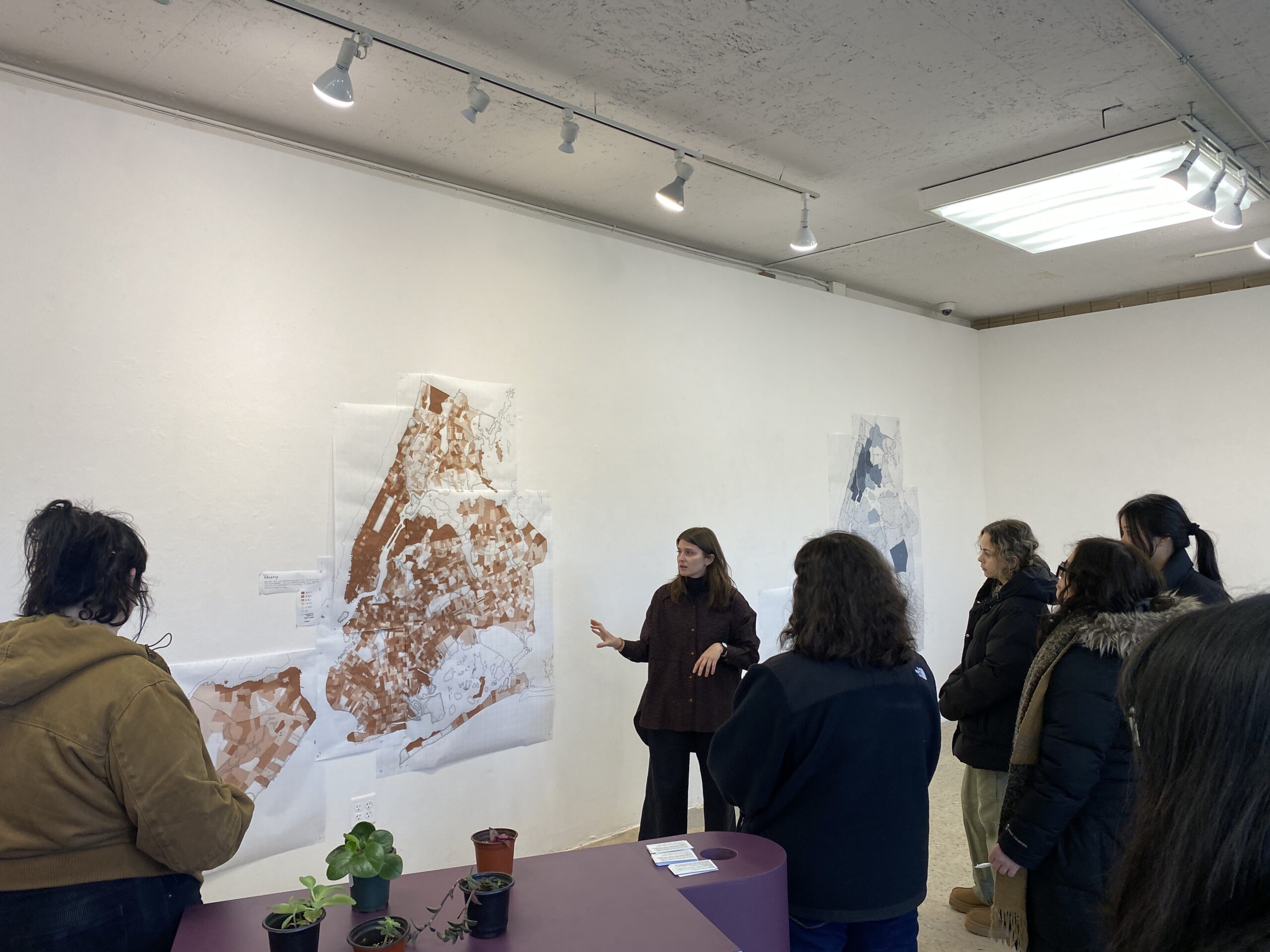 A group of people gather in an art gallery, listening to a speaker presenting detailed maps mounted on the wall. The maps show varying shades of brown and blue, indicating different areas or data. In the foreground, potted plants sit on a purple table. The audience appears engaged, some taking notes or looking closely at the maps while the speaker gestures toward them. The space has white walls and overhead lighting.
