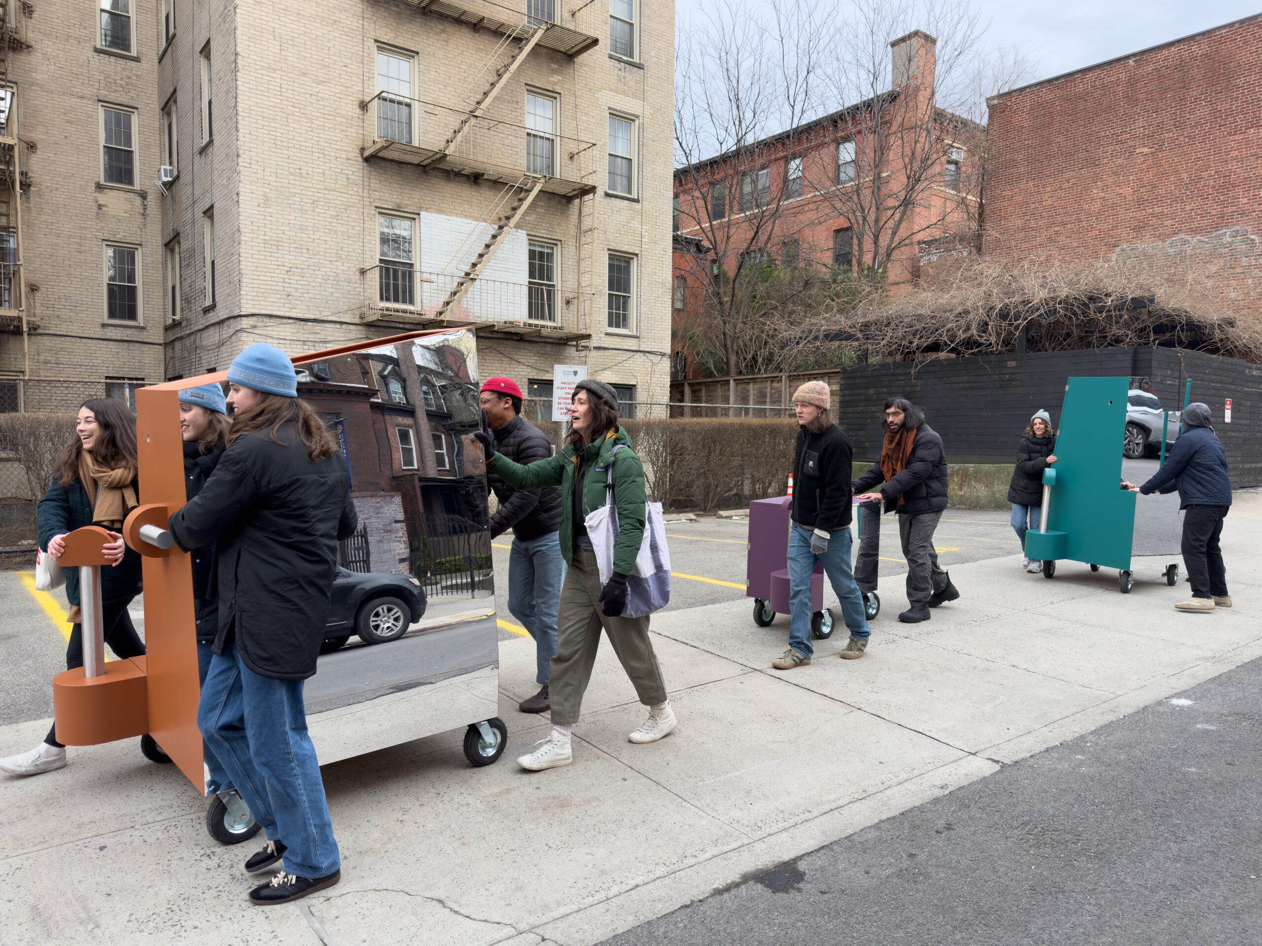 A group of seven people is moving colorful carts on a city street, with some carts featuring mirrors. They are dressed in winter clothing, including hats and jackets, and are situated near brick buildings. One cart has a reflective surface showing a nearby house, while others are solid colors (purple, green, and orange). The atmosphere appears casual and collaborative.