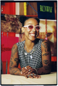 A person with hair styled in two buns smiles while sitting at a table. They wear pink, oversized sunglasses and a black-and-white checkered top, adorned with various rings and necklaces. Their arms are covered in intricate tattoos, and colorful backgrounds with fabric and signs are visible behind them, creating a vibrant, artistic atmosphere.