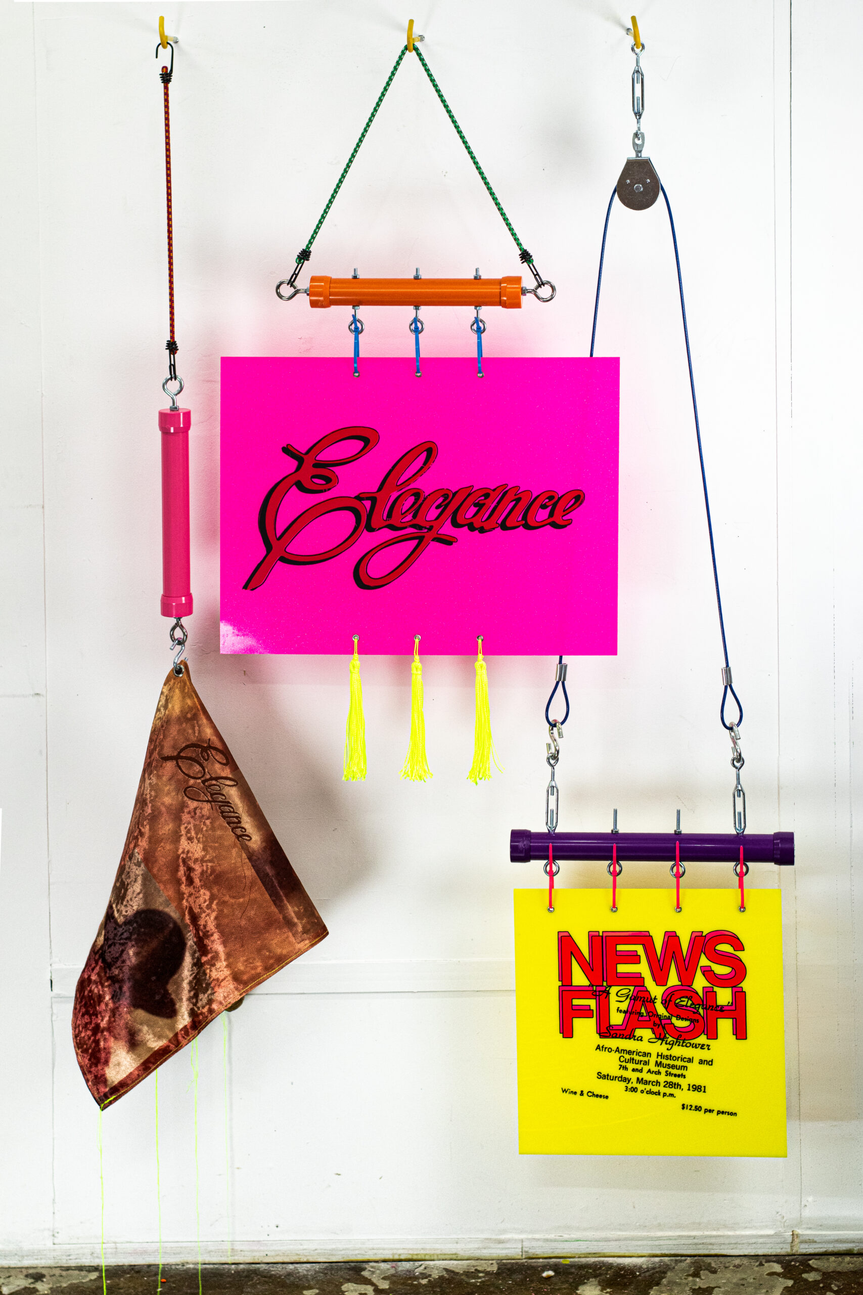A vibrant display of three hanging art pieces. The central piece is a bright pink sign with the word "Elegance" in cursive, complemented by yellow tassels. To the left hangs a triangular fabric with a brown and red design, also featuring the word "Elegance." The right side showcases a yellow sign that reads "NEWS FLASH" in bold red letters, attached with purple and silver hardware. The background is a plain white wall.