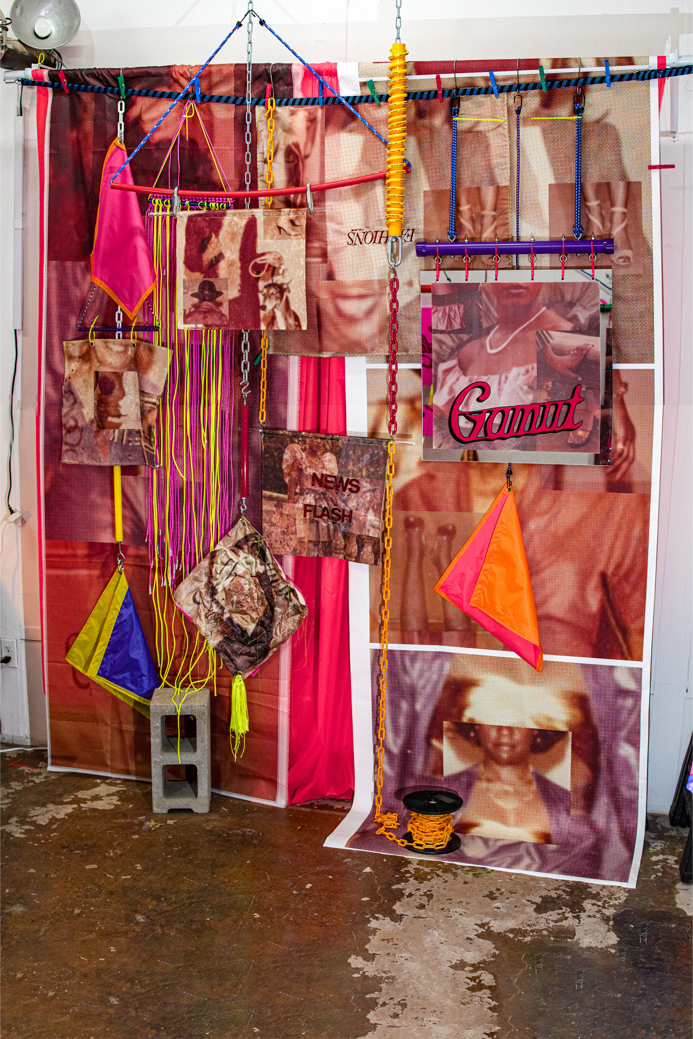 An artistic installation featuring a colorful backdrop composed of various fabric panels, including pinks and reds. Hanging from chains are several visually striking items, including printed fabric squares, bright flags, and textured materials. The focal point includes the word "Gamut" in bold lettering, along with other phrases like "NEWS FLASH." The ground has a stacked concrete block and a spool-like object. The overall composition combines vibrant colors and patterns, creating a dynamic visual display.