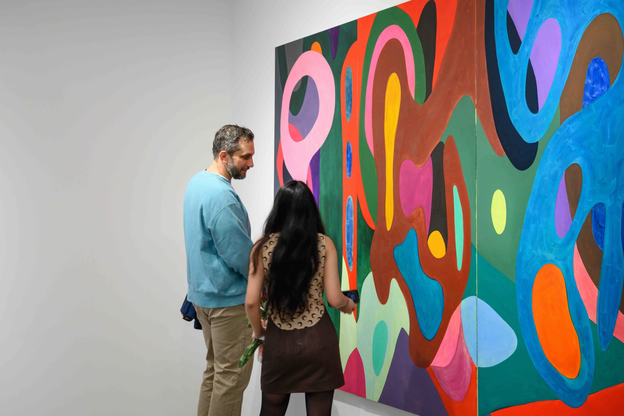 Two people are engaged in conversation while observing a colorful abstract painting on the wall. The artwork features various shapes and colors, including pinks, greens, blues, and reds. One person is facing the painting and holding a phone, while the other stands beside them, looking at the artwork and smiling. The setting appears to be an art gallery.