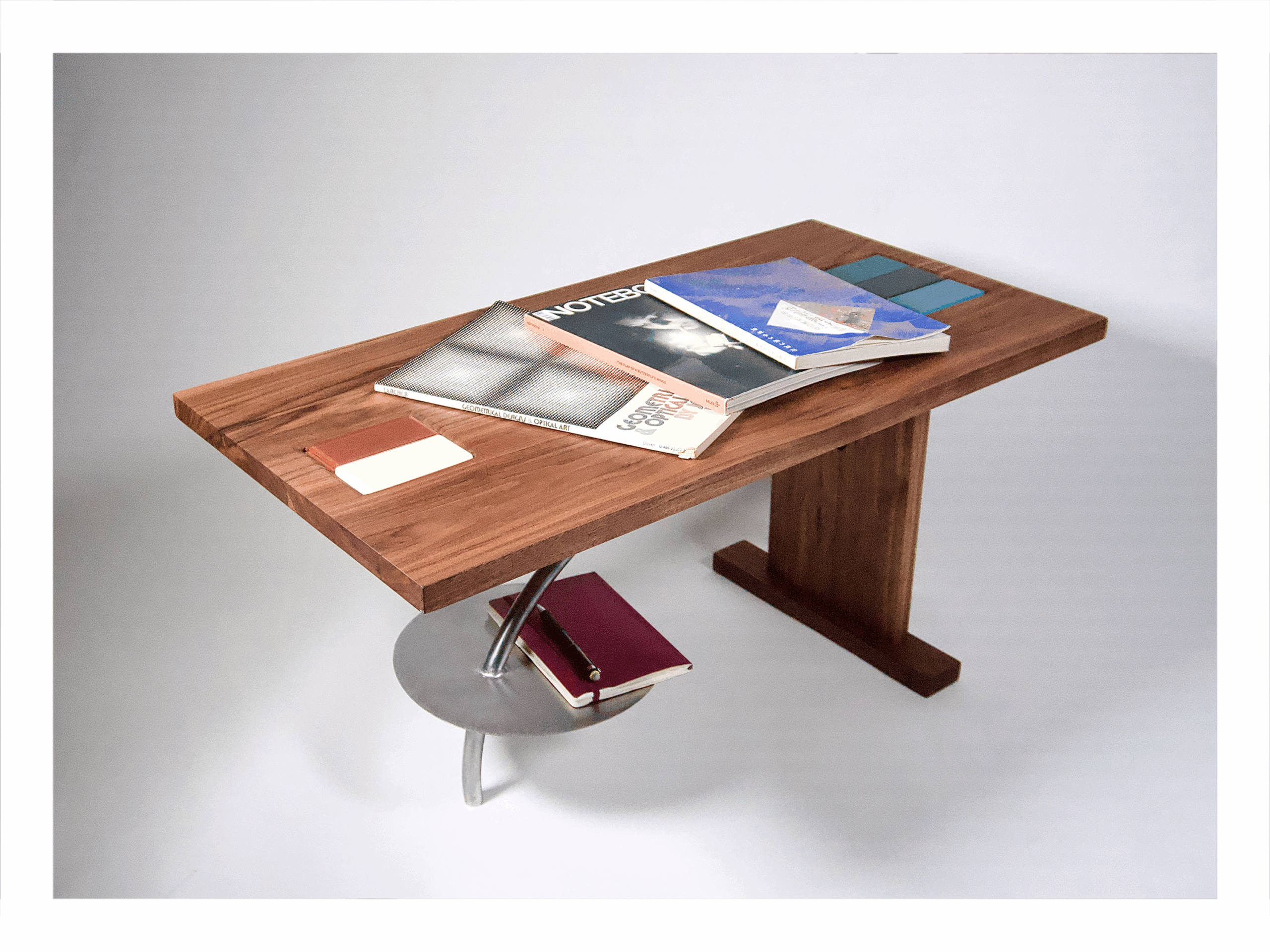 A wooden desk with a thick rectangular tabletop supported by a single wooden leg on one side and a curved metal support on the other. Various books, notebooks, and material samples are arranged on the surface. Beneath the tabletop, a circular metal shelf holds additional notebooks. The setup is photographed in a soft studio light against a neutral background.
