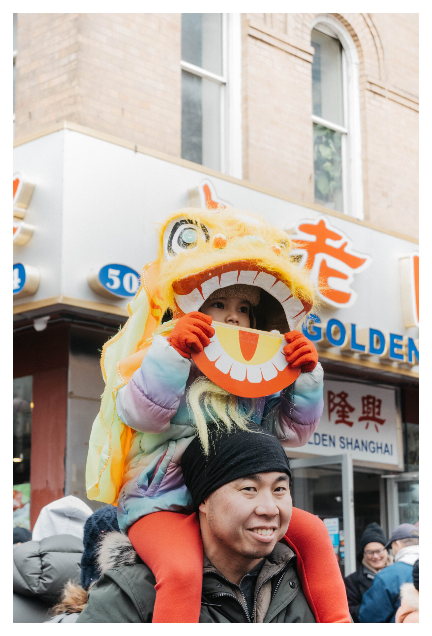 A photograph of a child sitting on an adult’s shoulders during a street celebration. The child wears a bright yellow lion-dance headpiece and holds a smiling lion-face cutout in front of their own face. The adult beneath is bundled in winter clothing and smiling. Behind them is a storefront with Chinese characters and the words “GOLDEN SHANGHAI.” People in winter jackets gather in the background.