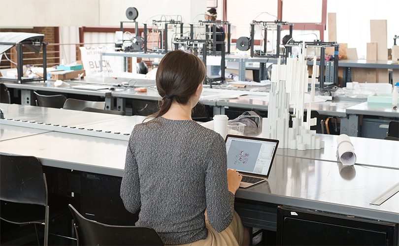A woman sits at a work table with a laptop. Sculptures and different tools and machinery are on the table with her.