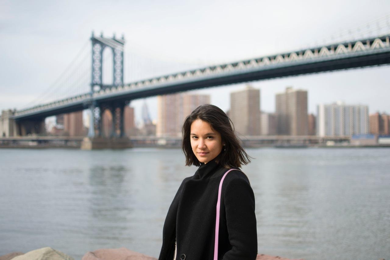 woman standing in front of manhattan bridge, with river behind her, woman wearing black coat and purse