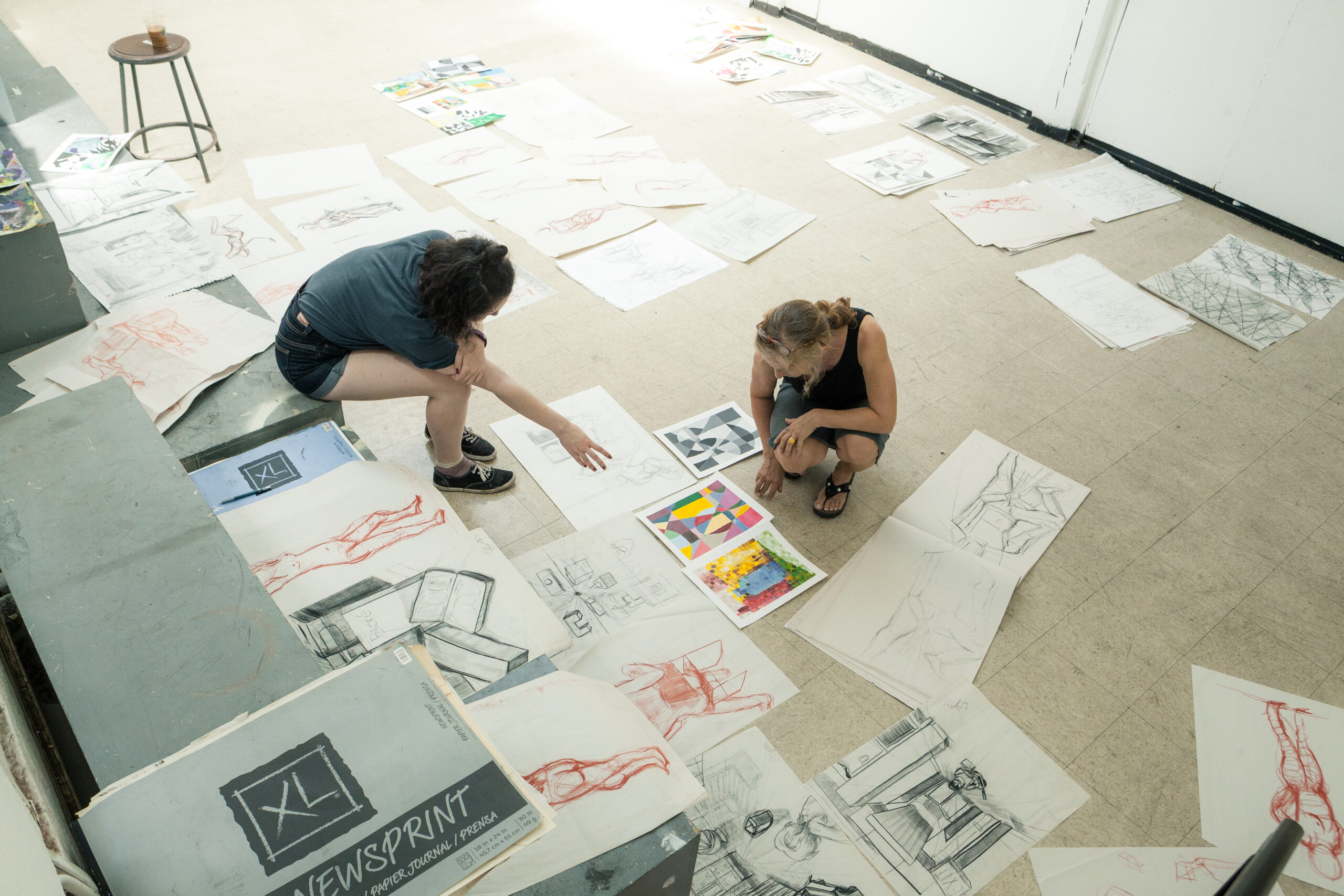 An art classroom scene where two individuals are kneeling on the floor, reviewing a collection of sketches and colorful artworks spread across the room. The drawings include figure studies, architectural sketches, and abstract designs. The workspace is casual, with newsprint pads and other materials scattered around, fostering a creative and exploratory environment.