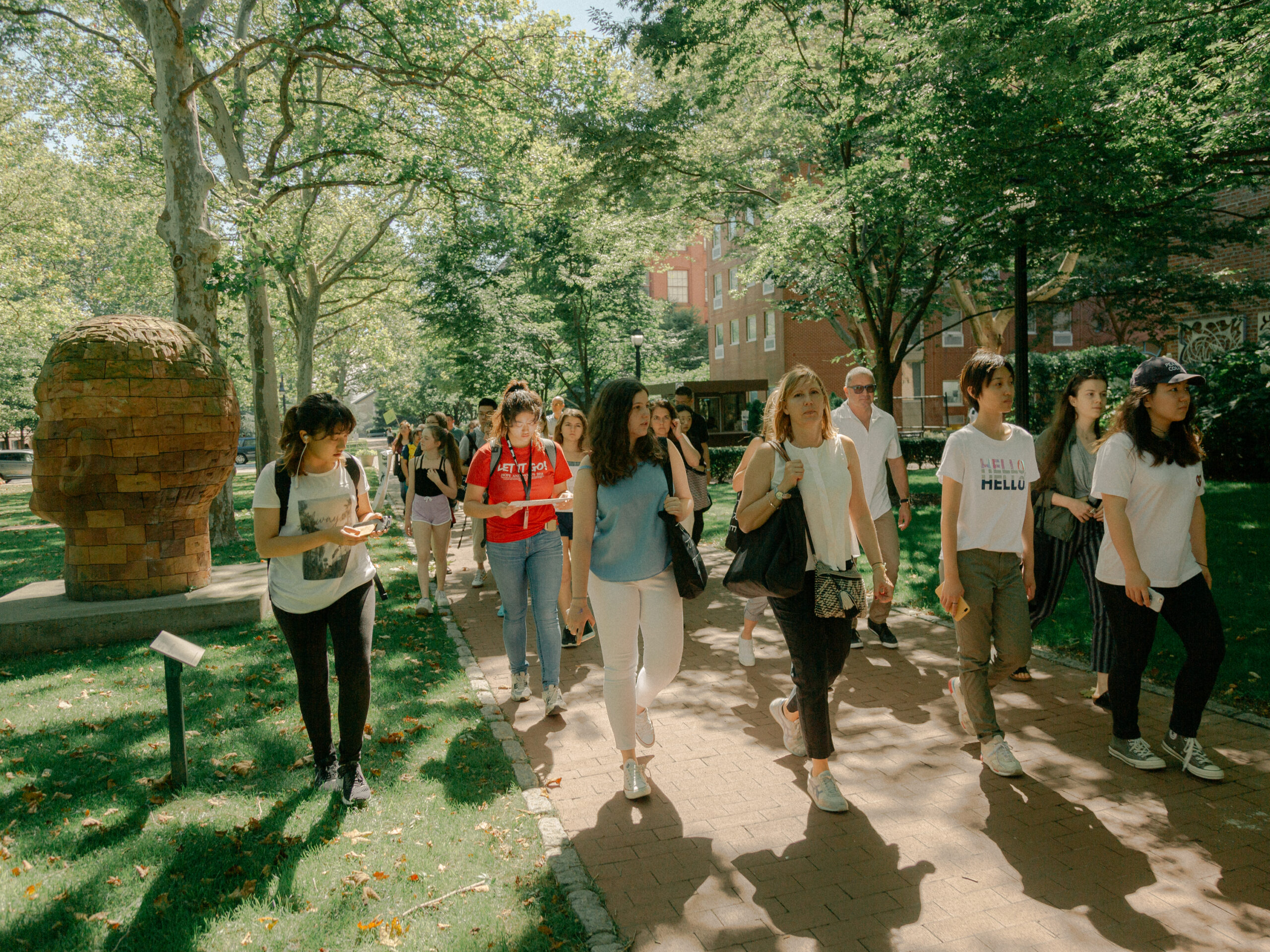 A group of students and parents walk through a tree-lined path on a college campus during a sunny day, likely a campus tour. The setting is lush and green, with dappled sunlight on the brick path and grass. A large brick sculpture of a head is visible on the left side of the image. Participants are casually dressed, with some carrying backpacks, phones, or bags. A red brick building and outdoor artwork are visible in the background.