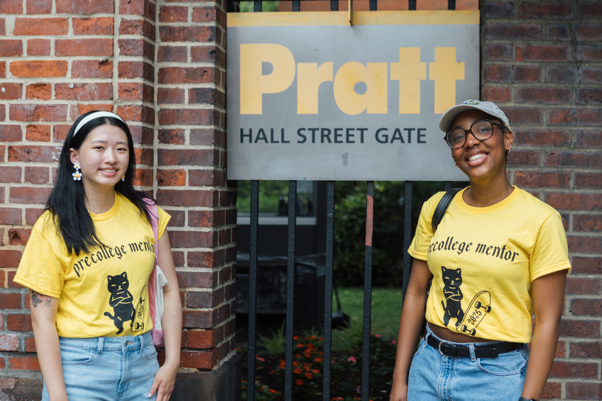 Two young women stand in front of the Pratt Hall Street Gate entrance sign, smiling at the camera. Both wear bright yellow "precollege mentor" T-shirts with a black cat graphic and denim bottoms. The woman on the left has long black hair, flower-shaped earrings, and a headband, while the woman on the right wears glasses, a baseball cap, and a backpack. The background shows a brick wall, metal gate, and greenery.