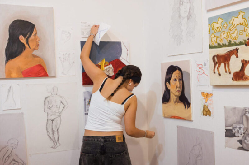 A student wearing a white tank top and dark jeans is arranging artwork on a white gallery wall. The person faces away from the camera, with their arm raised to pin or adjust a drawing. The wall is covered with various studies and paintings, including portrait paintings of women, figure drawings, hand and foot sketches, and a painting of foals with symbolic figures above them. The environment suggests an art classroom or exhibition setup displaying works in progress and completed pieces.