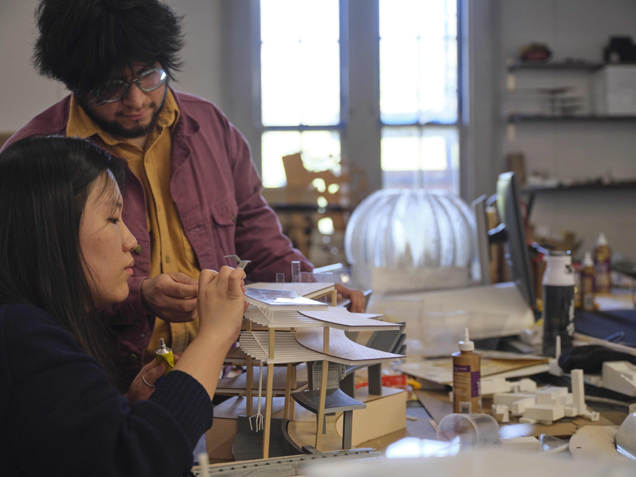 Two students work closely on a detailed architectural model in a studio setting. One student is applying glue to a small piece while the other observes and assists. The workspace is filled with supplies, tools, and additional models. Natural light enters through large windows in the background, illuminating the creative environment.