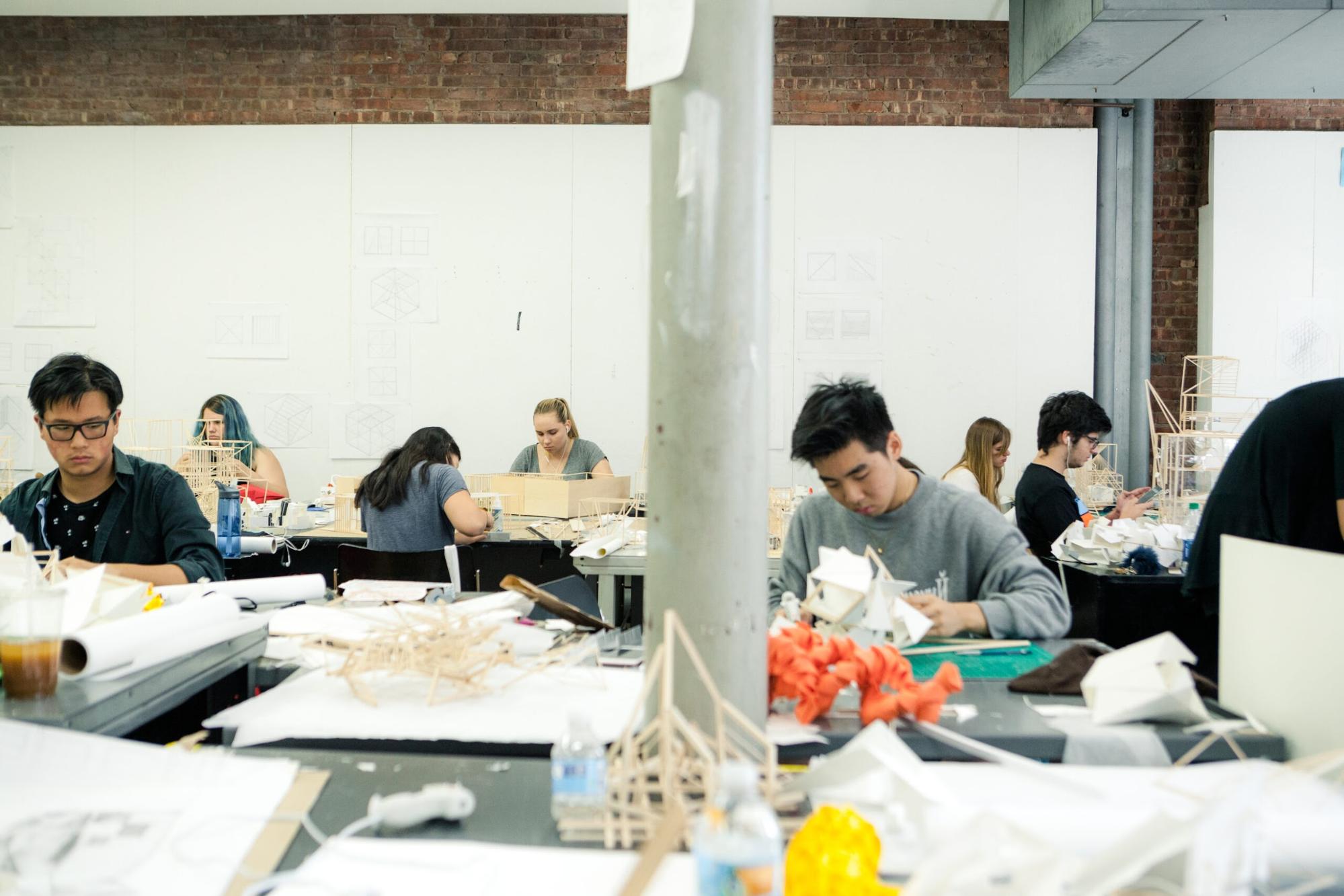 A classroom of architecture or design students working at tables filled with models, sketches, and materials. The students are focused on constructing small structures made of wood and paper, with drawings pinned on the wall behind them. The room has an industrial look with exposed brick and a central concrete column.