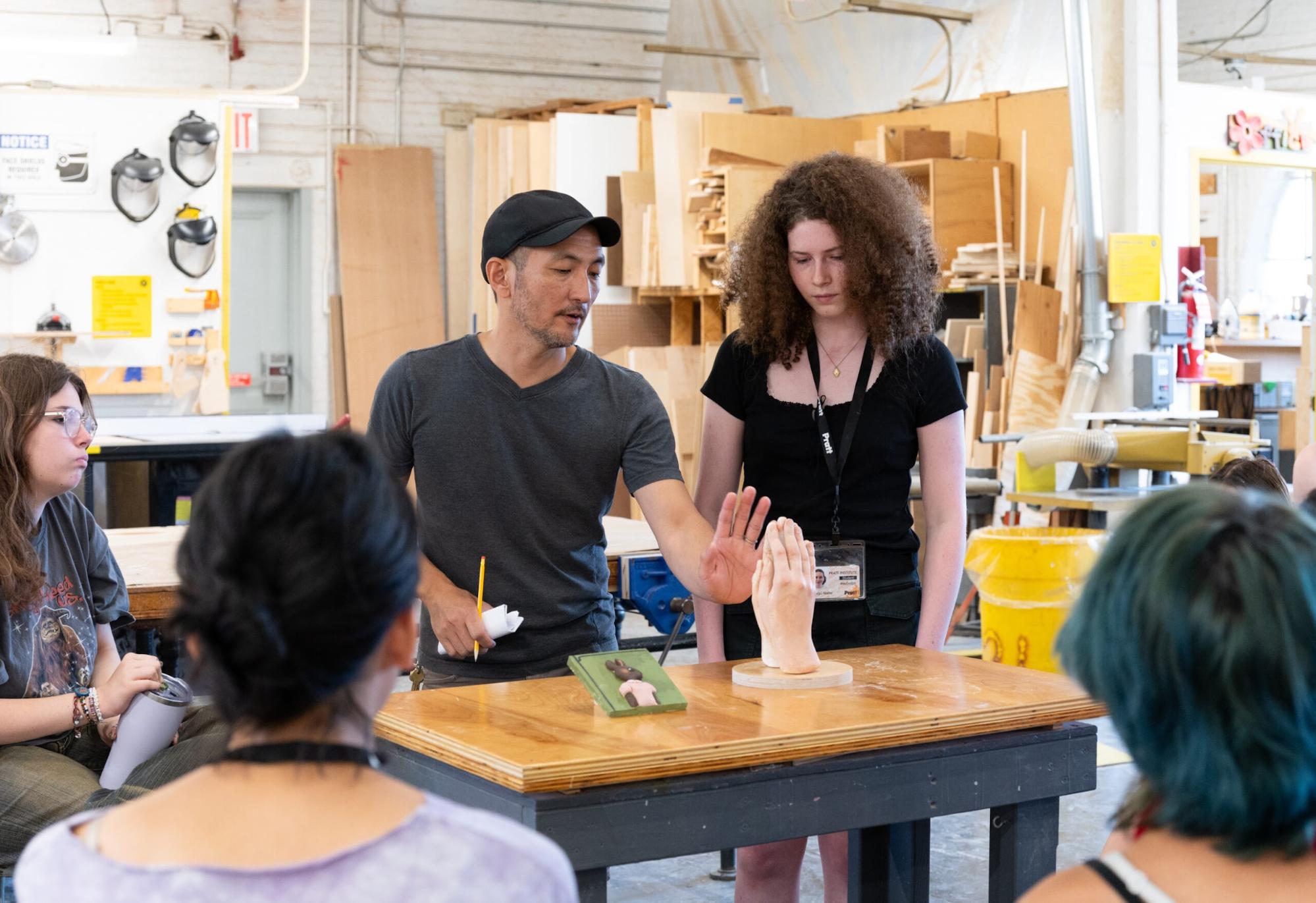 A classroom workshop scene where an instructor in a black cap and gray shirt is demonstrating to students. He gestures toward a realistic hand sculpture placed on a wooden table, alongside another model. A student with curly hair and a black shirt stands next to him, observing closely, while other students sit around the table watching. The background shows a woodworking studio with safety gear, tools, and wooden boards.