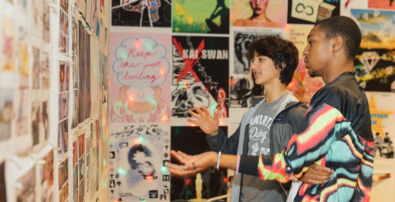 Two students stand indoors, looking at a wall covered with colorful posters and printed images. They appear to be discussing the display, gesturing with their hands. The posters feature text, graphics, and artwork in a collage-like arrangement.