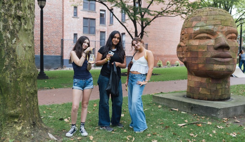 Three young women stand outdoors on a grassy area near a large sculptural head. One holds a drink, another smiles with her hand on her hip, and the third poses casually. Behind them is a brick building and trees, suggesting a campus or park setting.