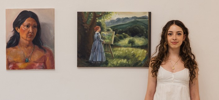 A young woman with long curly hair stands in front of two framed paintings on a gallery wall. One painting depicts a woman in a portrait style, and the other shows a figure painting outdoors in a landscape. The student smiles, suggesting she may be the artist presenting her work.