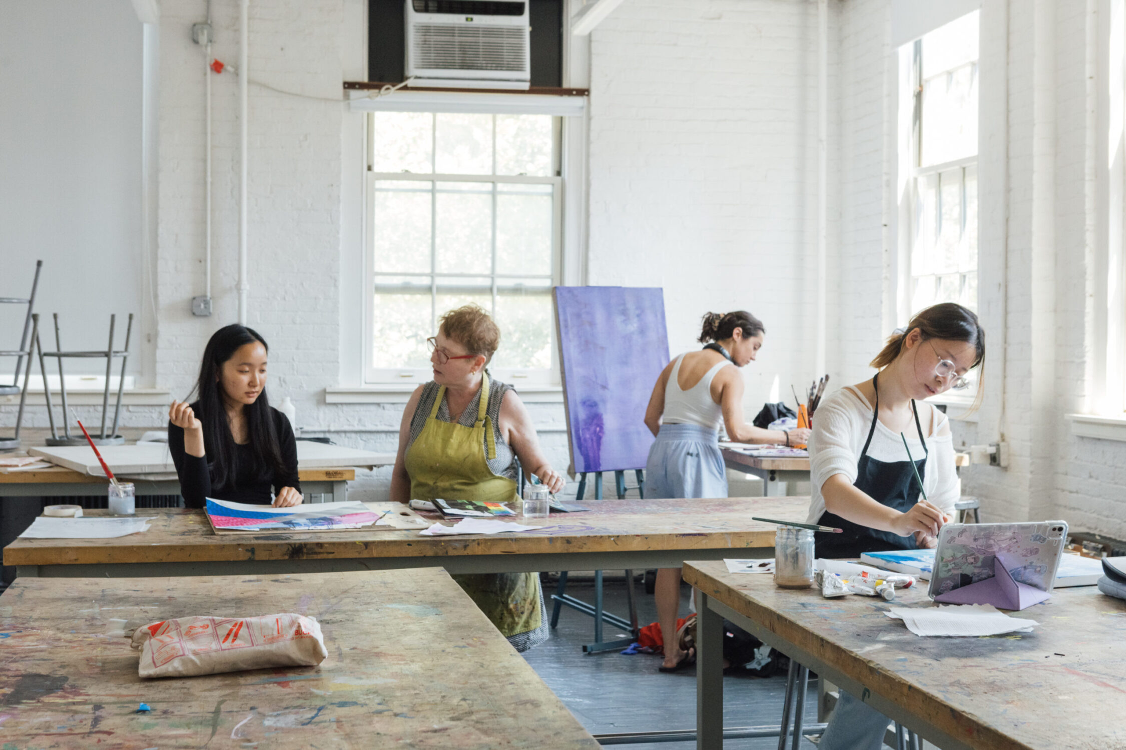 Inside a spacious art studio, three women are engaged in creative activities. Two women are seated at a wooden table, discussing and working on colorful paper art, while a third woman stands nearby, painting on a canvas. The room features large windows that allow natural light to fill the space, highlighting the artistic environment filled with supplies and artwork.