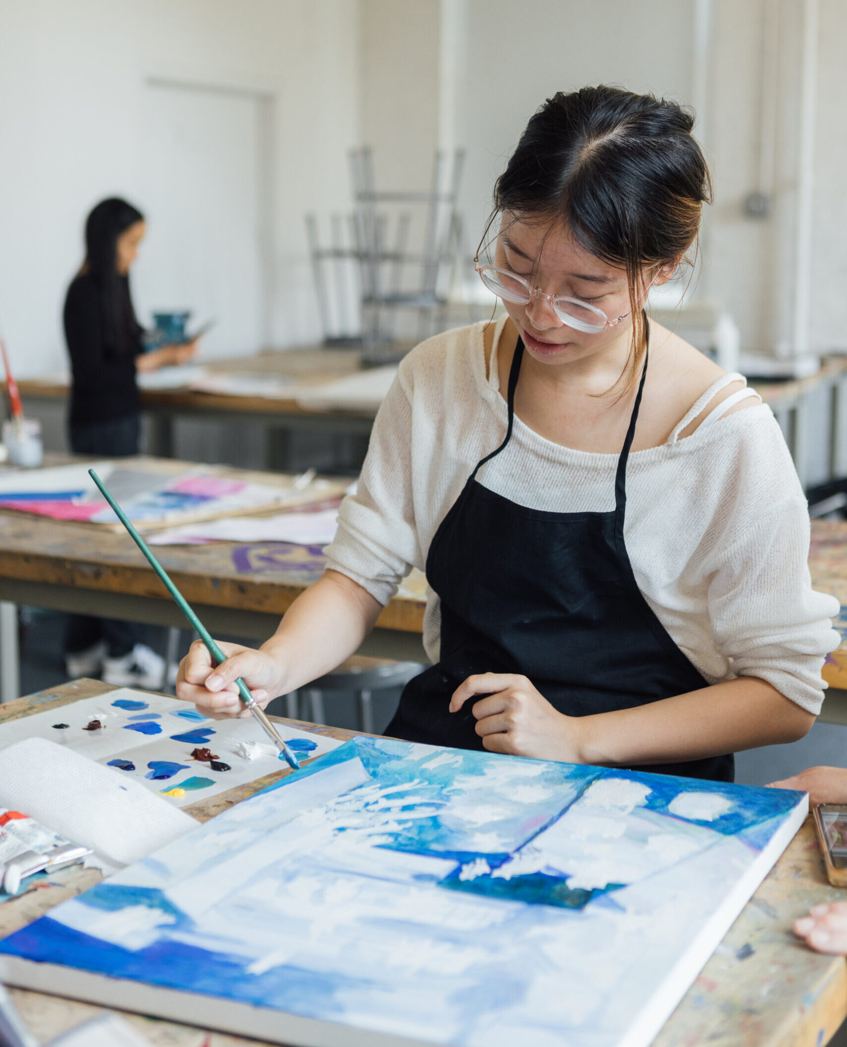 A woman wearing glasses and a black apron paints on a canvas in a bright art studio. She focuses intently on her work, using a green paintbrush to add blue tones to her artwork. In the background, another person stands at a table, engaged in a different creative task. Various art supplies, including paint tubes and palettes, are scattered on the table.