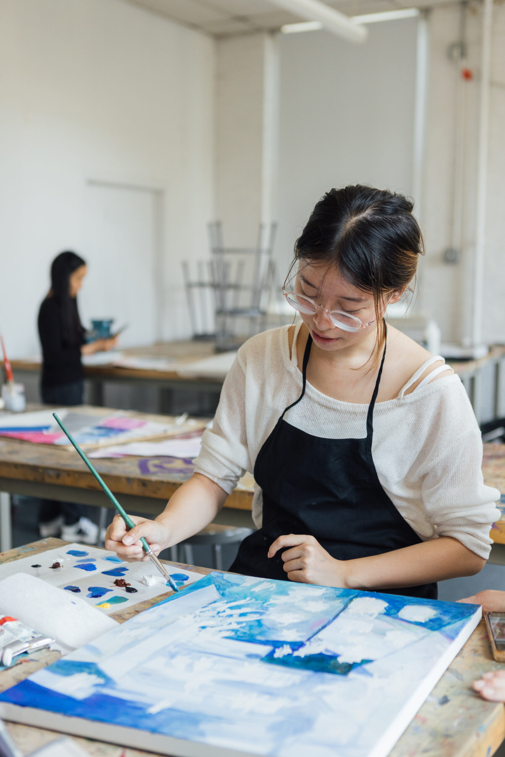 A woman wearing glasses and a black apron paints on a canvas in a bright art studio. She focuses intently on her work, using a green paintbrush to add blue tones to her artwork. In the background, another person stands at a table, engaged in a different creative task. Various art supplies, including paint tubes and palettes, are scattered on the table.