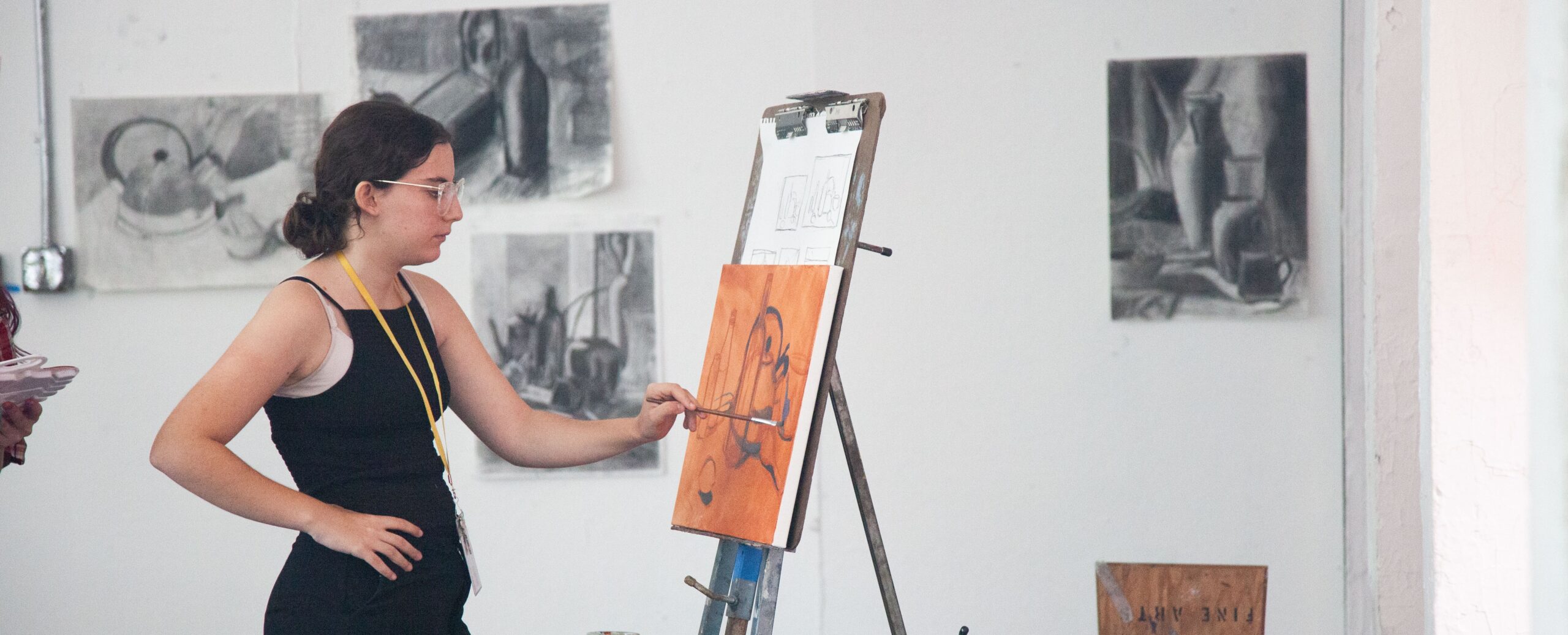 A student in a drawing studio stands at an easel, applying paint to an orange underpainting of a still-life scene. Charcoal still-life drawings are displayed on the wall behind them. The student wears glasses and a yellow lanyard and appears focused as they paint.