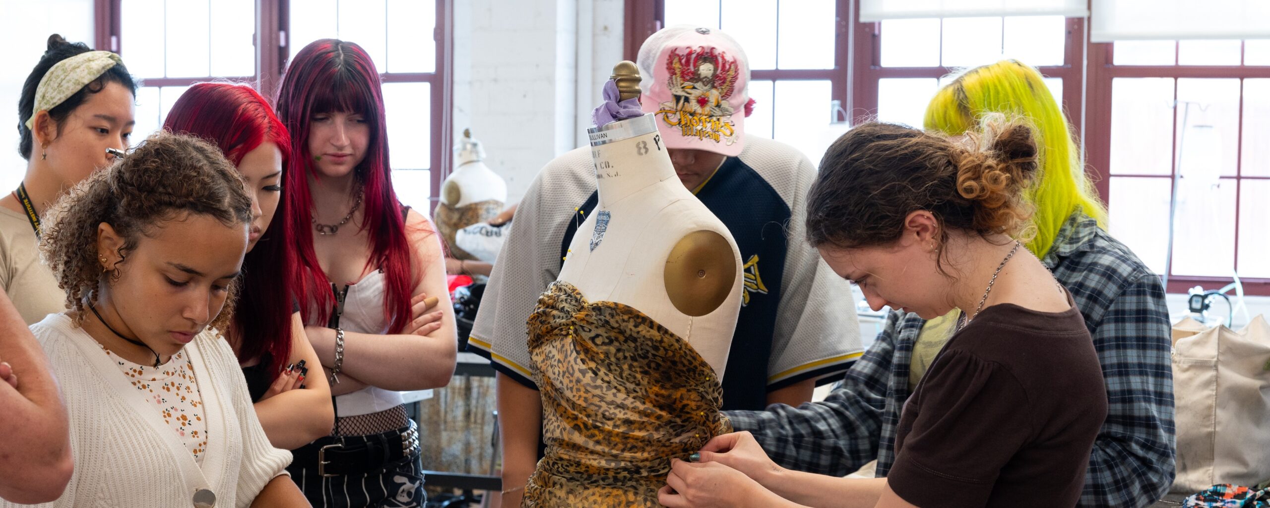 A group of three students, in a well-lit classroom, work around a mannequin form, working with fabric textiles