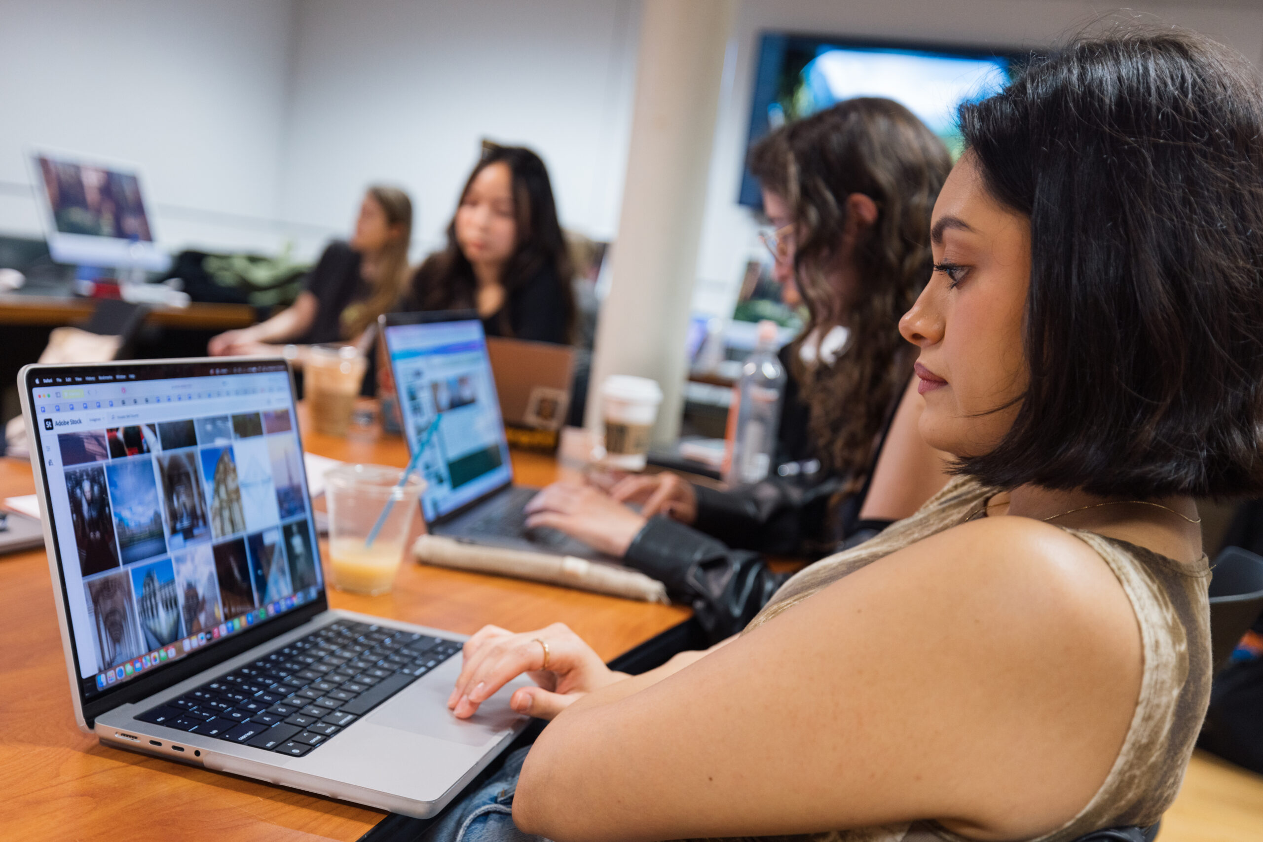 A group of people is seated around a table in a workspace, focused on their laptops. In the foreground, a woman with short dark hair is using a laptop displaying a grid of images. Other individuals are partially visible in the background, also engaged with their devices. One person has a coffee cup, and there are some drinks on the table. The atmosphere is collaborative and tech-oriented.
