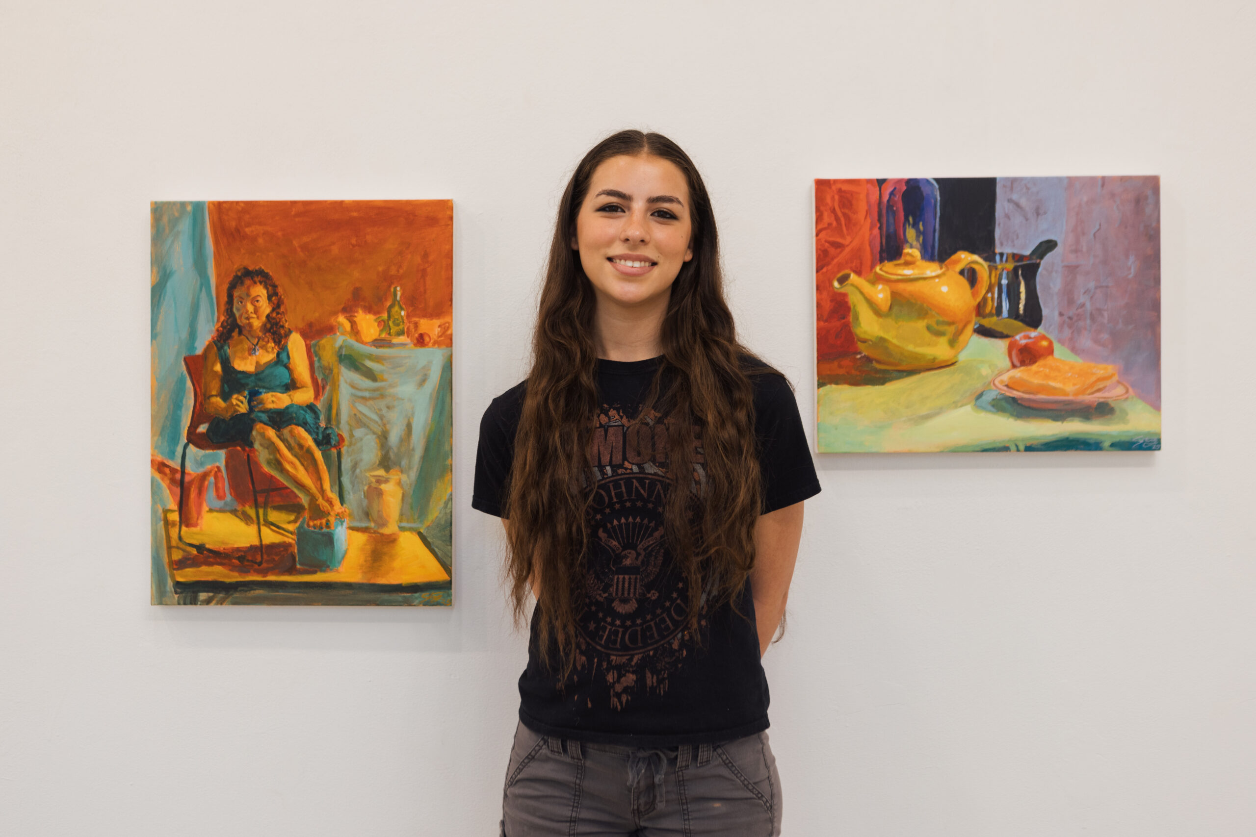 A young woman with long, wavy brown hair stands smiling in front of two colorful paintings on a white wall. To her left is a portrait of a woman sitting in a chair, dressed in a blue outfit. To her right is a still life featuring a yellow teapot, an apple, and a plate on a green tablecloth. The setting suggests an art gallery or exhibition.