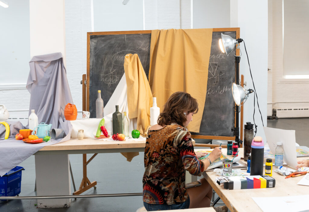 A young woman sits at a table, painting on a canvas in an artist studio. The table is cluttered with various art supplies, including paint bottles and paper. In the background, a black chalkboard is partially covered with fabric drapes, and colorful still-life objects, such as fruits and pottery, are arranged on the table. The scene is well-lit by lamps and the natural light from a nearby window.