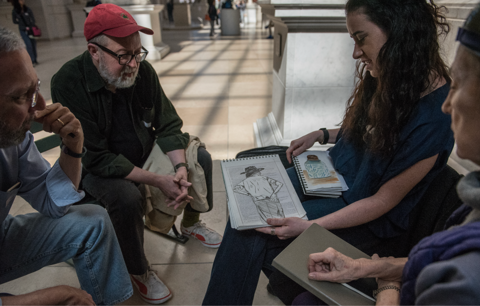 A group of four people is seated in a well-lit space, engaged in a discussion. One person is showing a sketchbook with an illustrated figure to the others. The individuals are dressed casually, with some wearing glasses and a baseball cap. The background features architectural details and people walking in the distance, conveying a lively atmosphere.